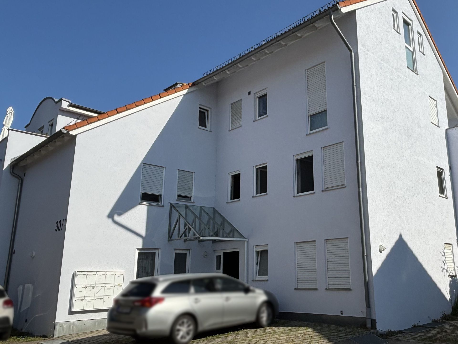 Das Bild zeigt ein Mehrfamilienhaus mit weißer Fassade und einem Vordach über dem Eingangsbereich.  Vor dem Haus befindet sich ein gepflasterter Bereich mit Briefkästen und ein Auto parkt davor.