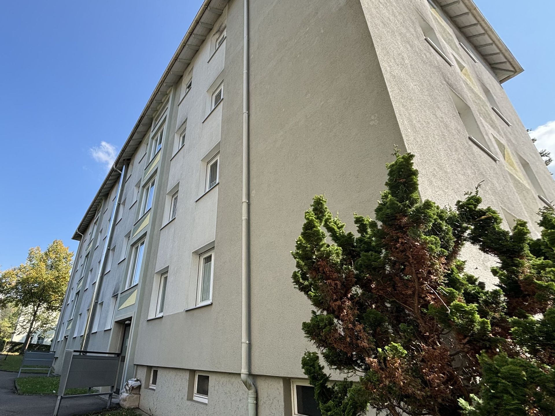 Das Bild zeigt ein Mehrfamilienhaus mit heller Fassade und einer Reihe von gelben und grünen geometrischen Mustern, die zwischen den Fenstern angebracht sind. Vor dem Haus befindet sich ein grüner Busch.