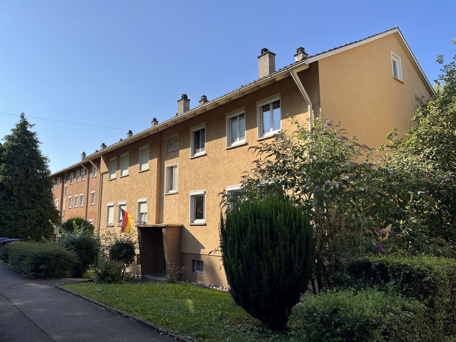 Das Bild zeigt ein Mehrfamilienhaus mit hellbrauner Fassade und weißen Fensterrahmen. Vor den Häusern erstreckt sich ein kleiner Vorgarten mit Rasenfläche, Büschen und Bäumen.