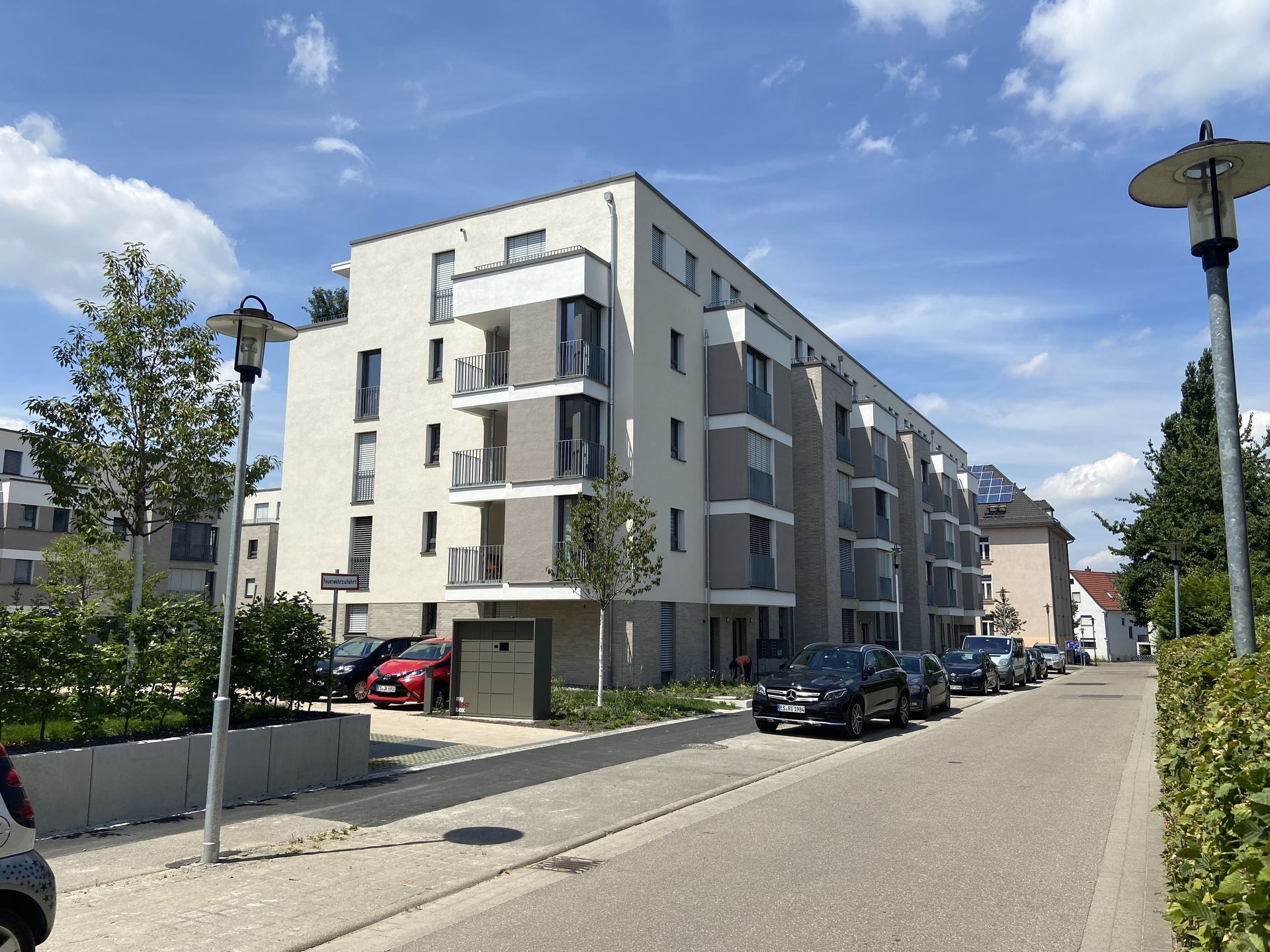 Das Bild zeigt ein Wohnhaus mit weißer Fassade und grauen Balkonen. Vor dem Haus sind Autos geparkt.