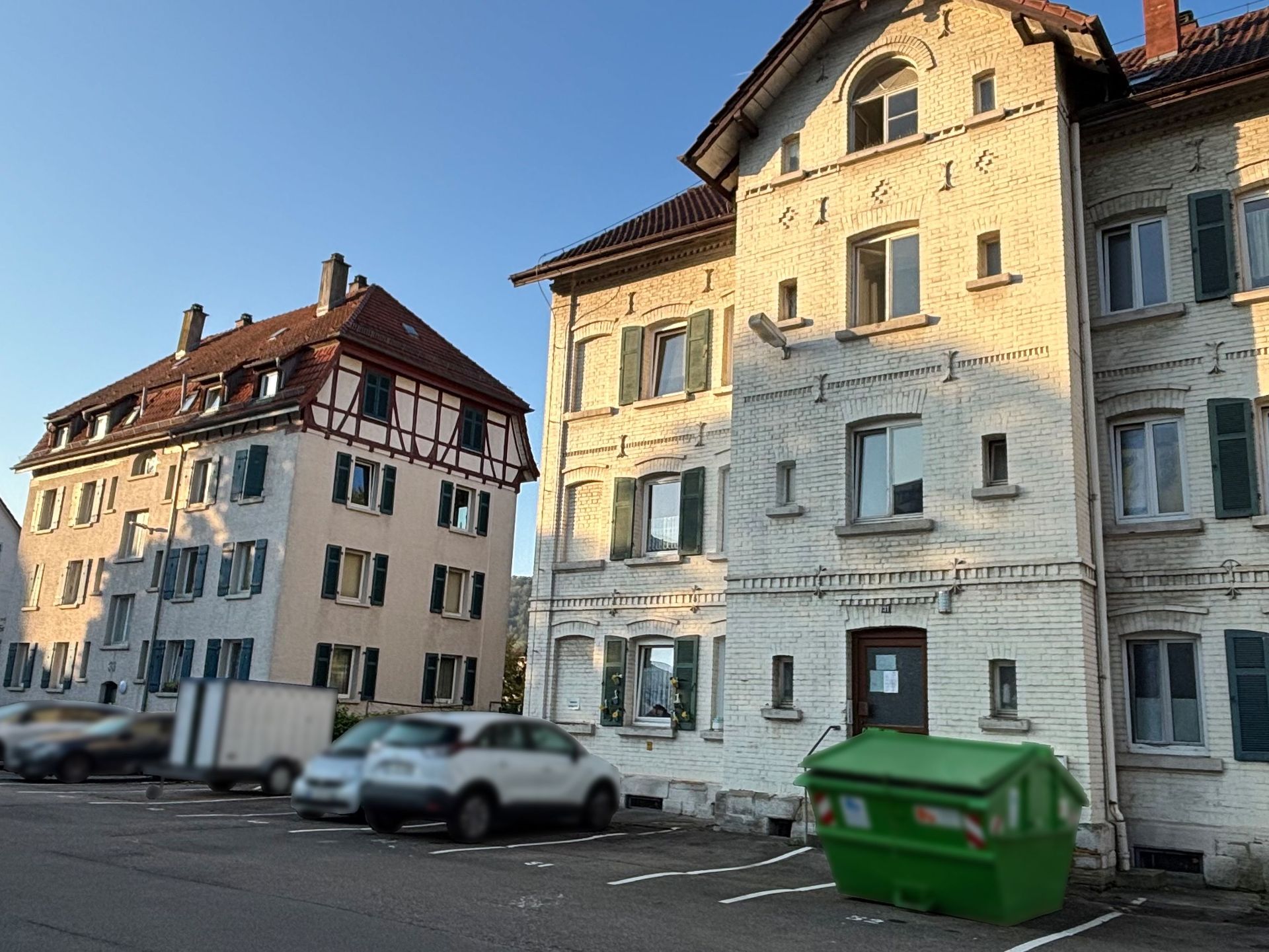 Das Bild zeigt zwei Mehrfamilienhäuser mit weißer Fassade und grünen Fensterläden. Vor den Häusern befindet sich ein Parkplatz mit mehreren Autos.
