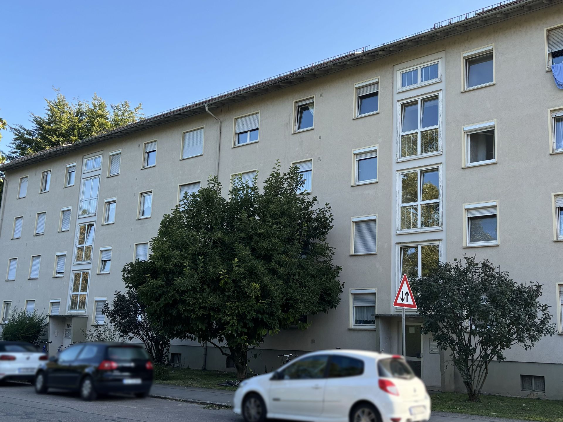 Das Bild zeigt ein Mehrfamilienhaus mit heller Fassade und vielen Fenstern. Vor dem Haus stehen Bäume und parkende Autos.
