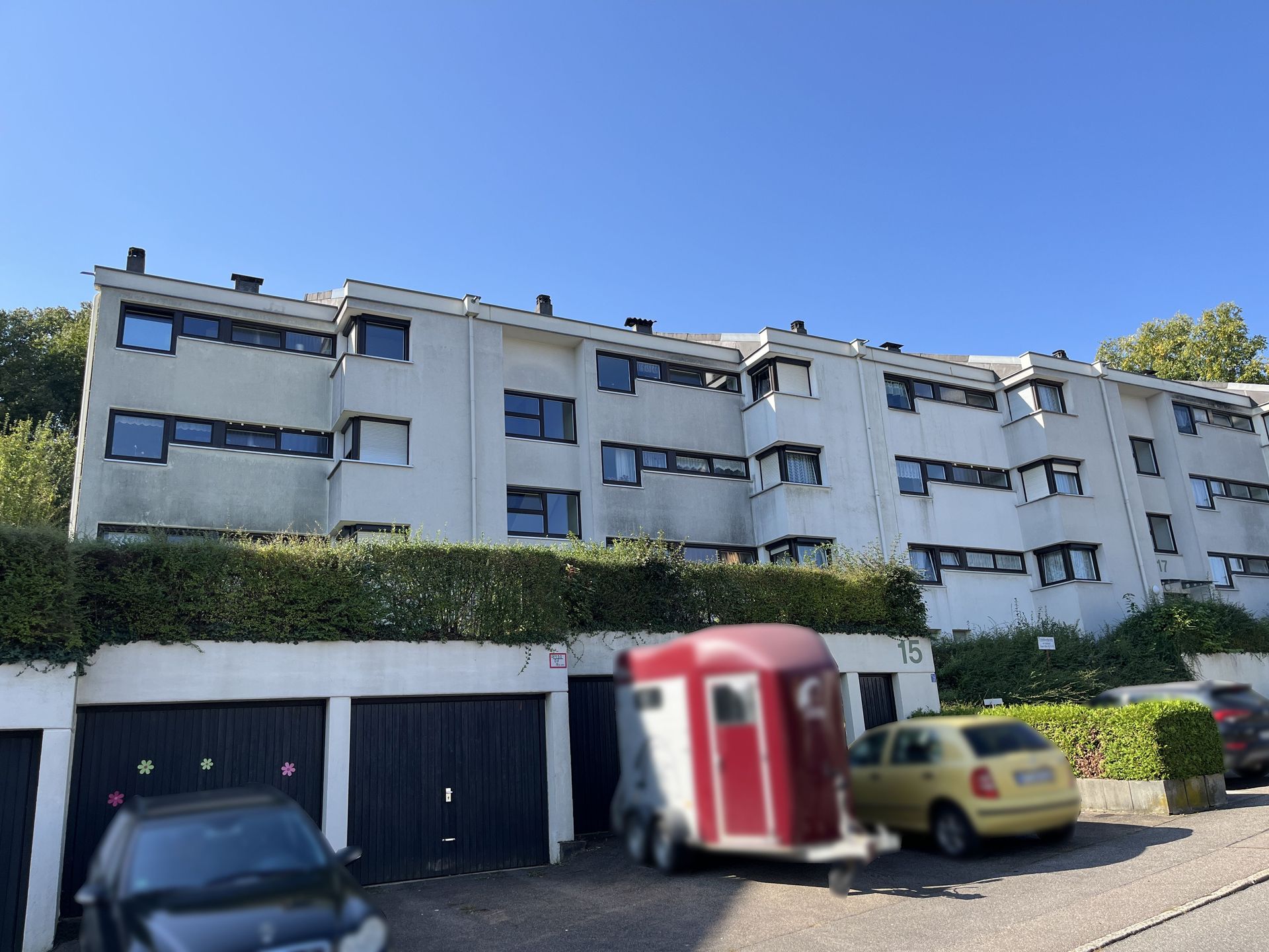 Das Bild zeigt ein Mehrfamilienhaus mit heller Fassade und mehreren Stockwerken. Vor dem Haus befinden sich Garagen und Parkplätze, auf denen Autos und ein Pferdeanhänger abgestellt sind.