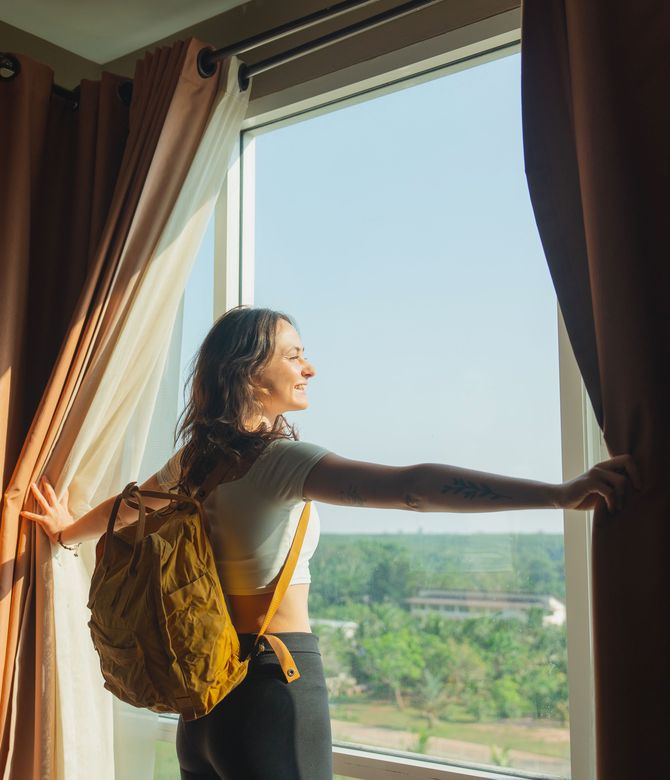 Das Bild zeigt eine Frau mit Rucksack, die aus einem Fenster schaut und die Gardinen zur Seite schiebt. Link zum Bereich Gästewohnung.
