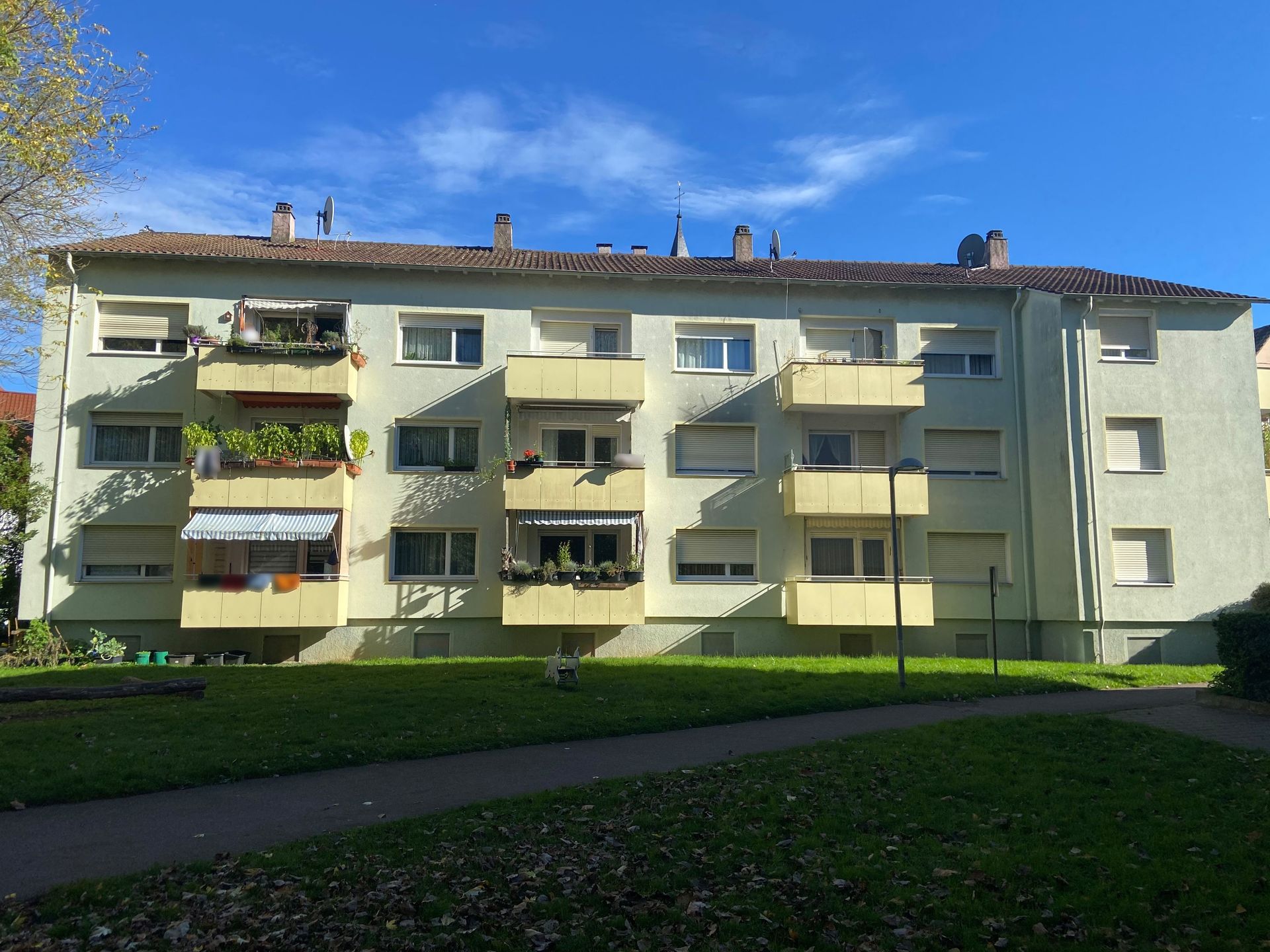 Das Bild zeigt ein Mehrfamilienhaus mit hellgrüner Fassade und gelben Balkonen, auf denen Pflanzen zu sehen sind. Vor dem Haus befindet sich eine große Rasenfläche.