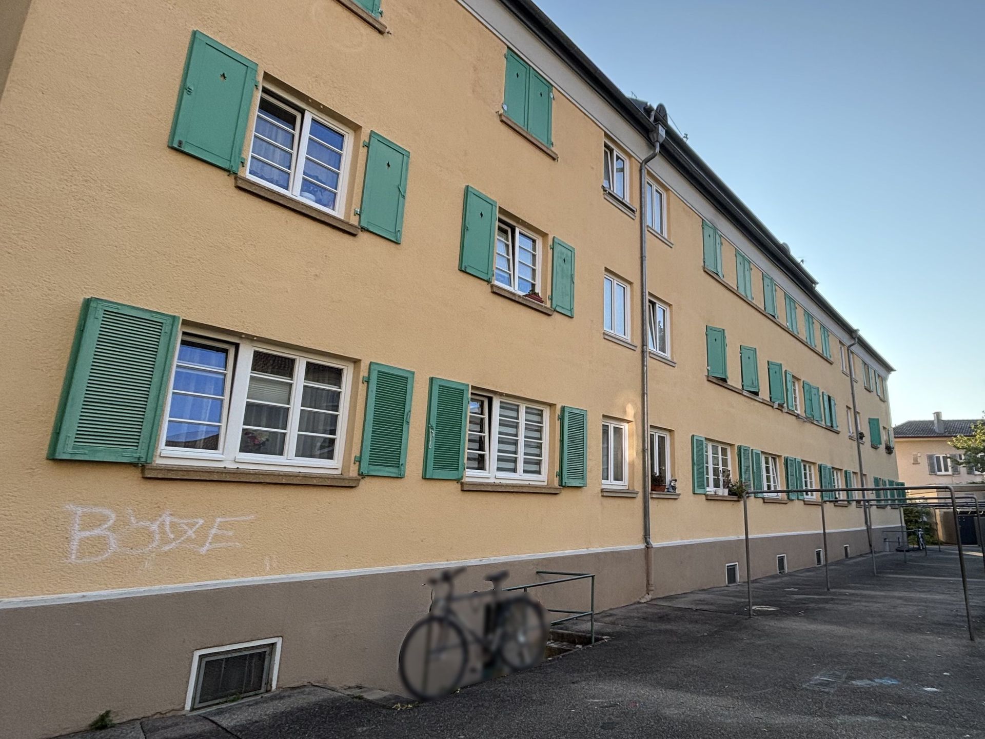 Das Bild zeigt ein Mehrfamilienhaus mit hellgelber Fassade und grünen Fensterläden.