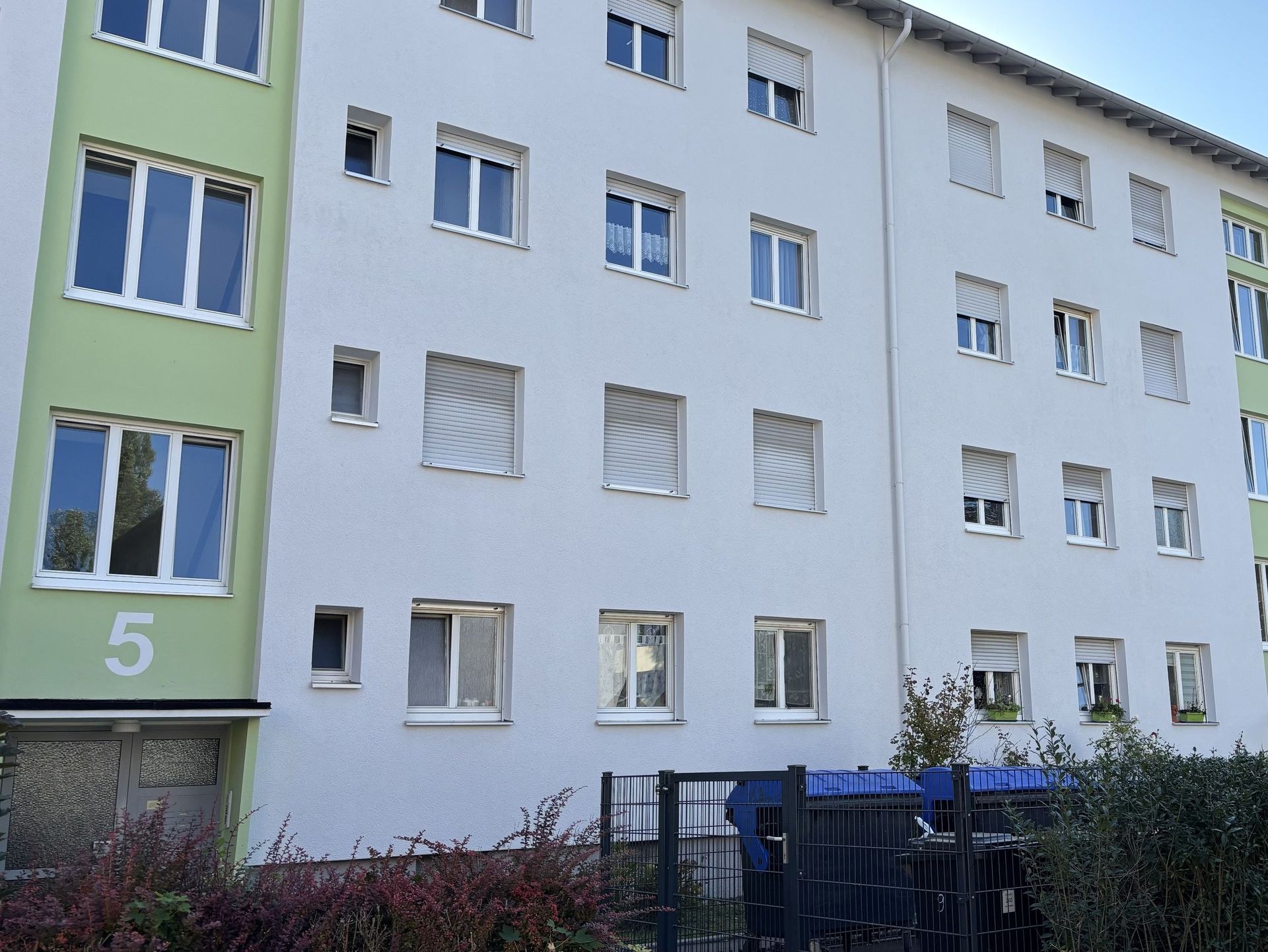 Das Bild zeigt ein Mehrfamilienhaus mit weißer Fassade und einigen hellgrünen Elementen. Vor dem Haus stehen Mülltonnen und ein kleiner Zaun begrenzt das Grundstück.