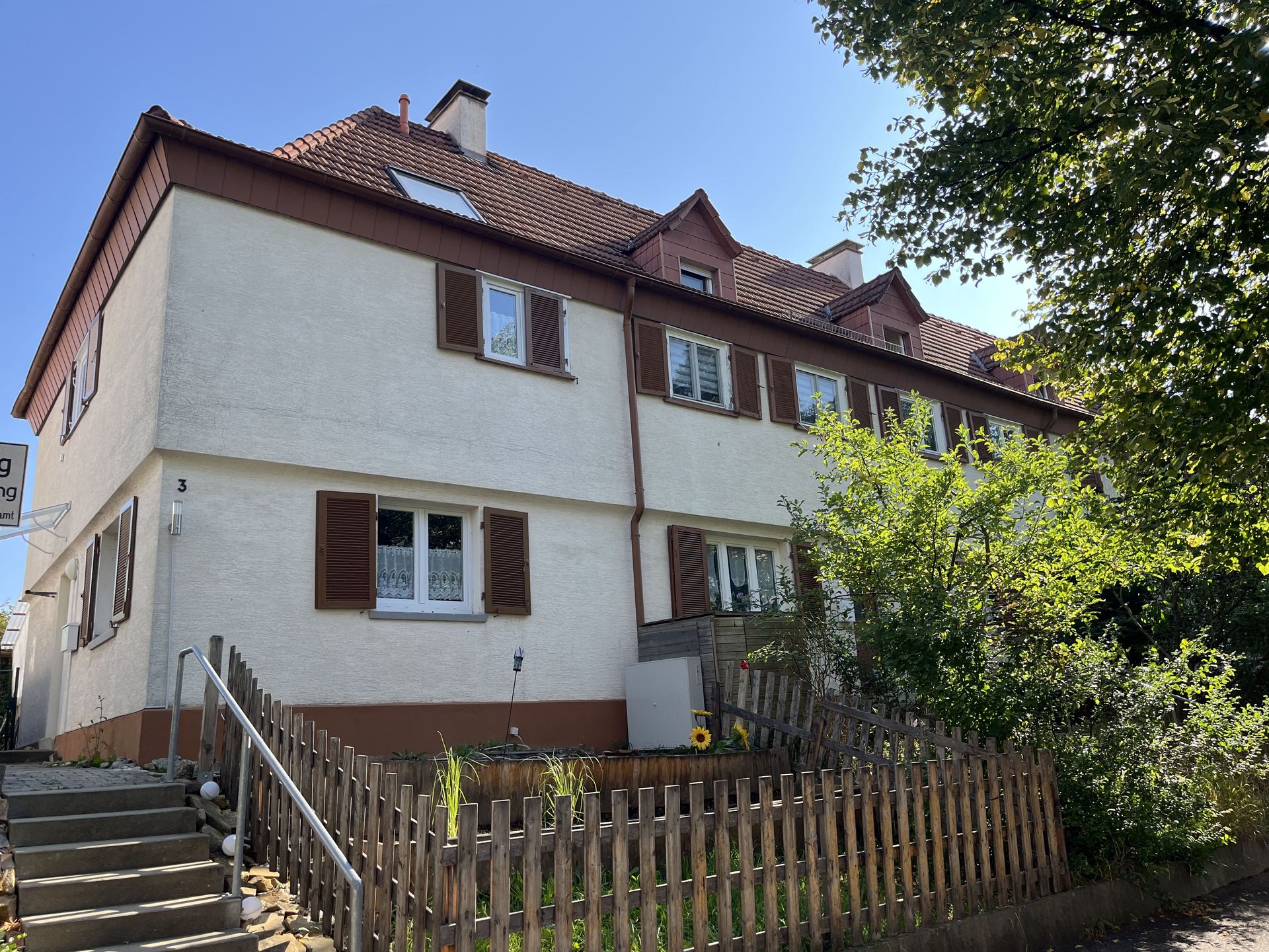 Das Bild zeigt ein Haus mit weißer Fassade, braunen Fensterläden und einem roten Ziegeldach. Vor den Häusern befindet sich ein kleiner Vorgarten mit einem Holzzaun und etwas Bepflanzung.
