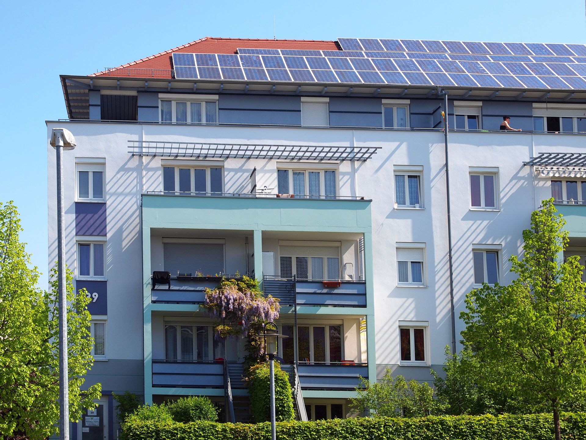 Das Bild zeigt ein modernes Mehrfamilienhaus mit weißer Fassade, das durch farbige Elemente an den Balkonen und Fensterbänken aufgelockert wird. Auf dem Dach des Gebäudes ist eine Solaranlage installiert und vor dem Haus stehen Bäume.
