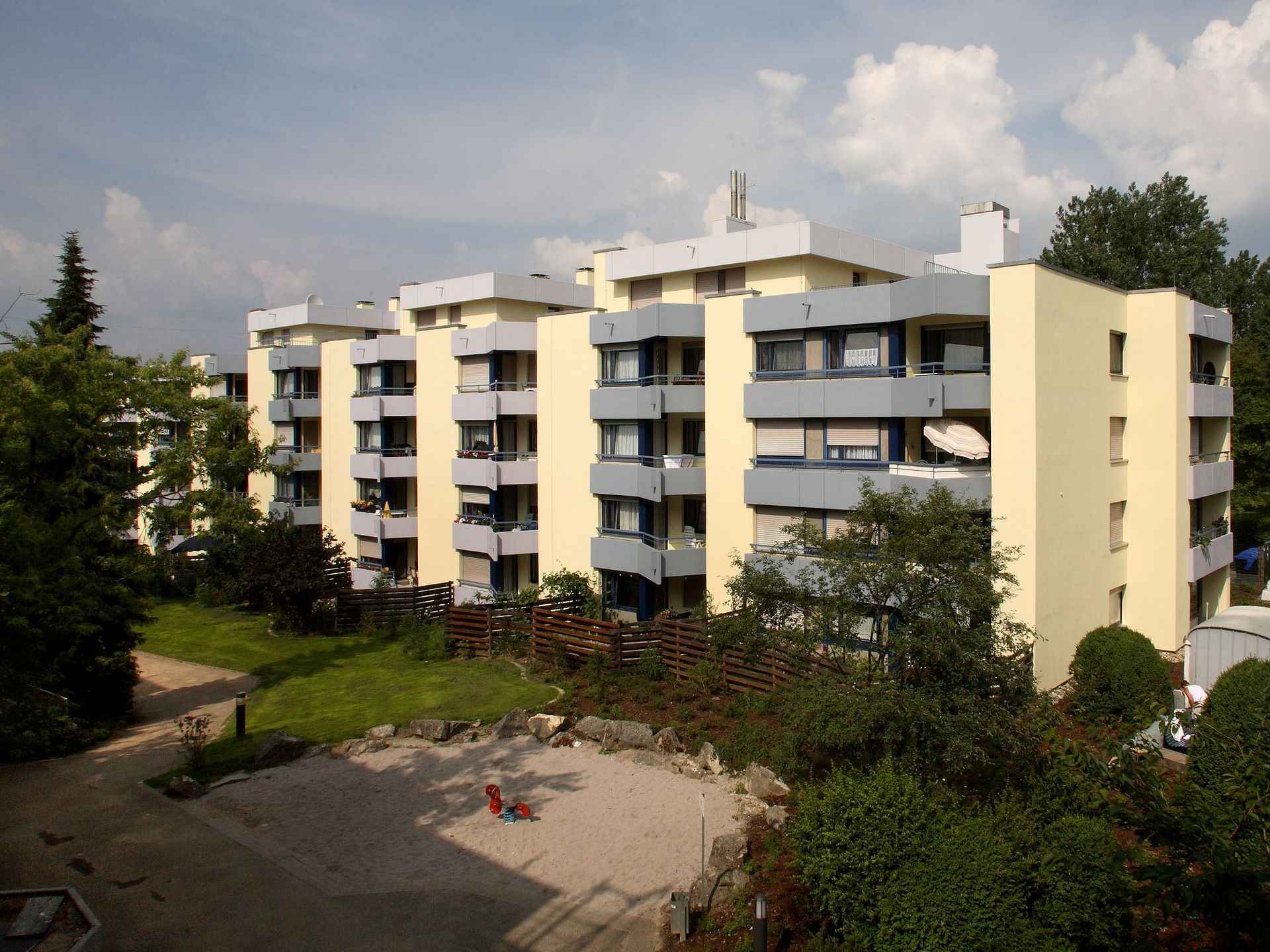 Die Wohnanlage besteht aus mehreren aneinandergereihten Mehrfamilienhäusern mit hellgelber Fassade und grauen Balkonen, die einen modernen und gepflegten Eindruck machen. Umgeben sind die Häuser von einer Grünfläche mit Bäumen, Sträuchern und einem Sandkasten im Vordergrund.