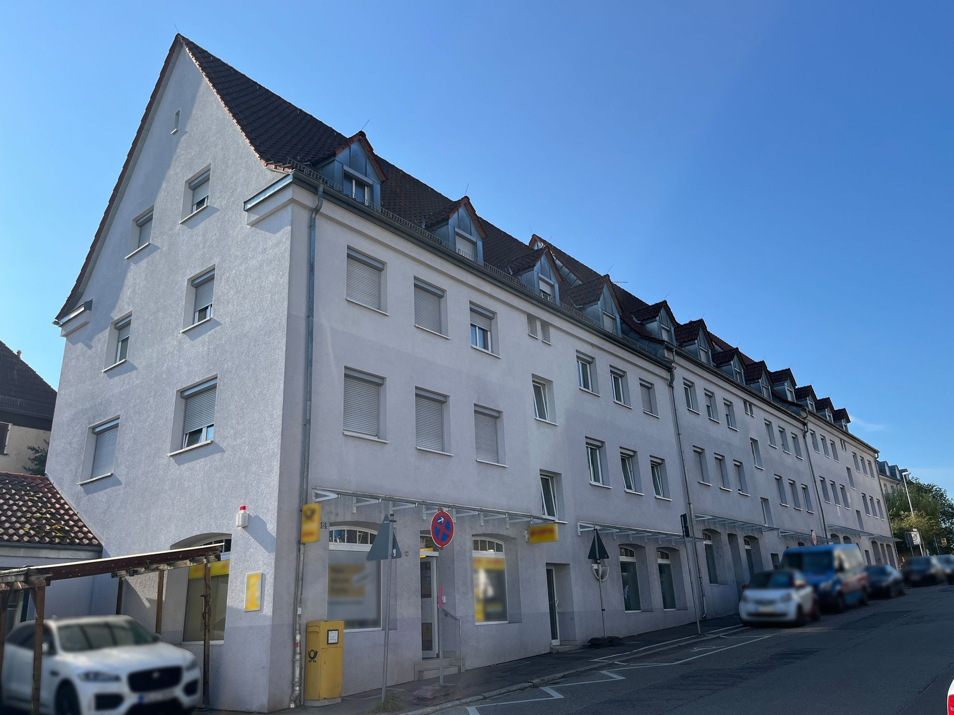 Das Bild zeigt ein Mehrfamilienhaus mit weißer Fassade, braunem Dach und mehreren Dachgauben. Im Erdgeschoss des Gebäudes befinden sich Ladenlokale, darunter eine Postfiliale, und davor parken Autos am Straßenrand.