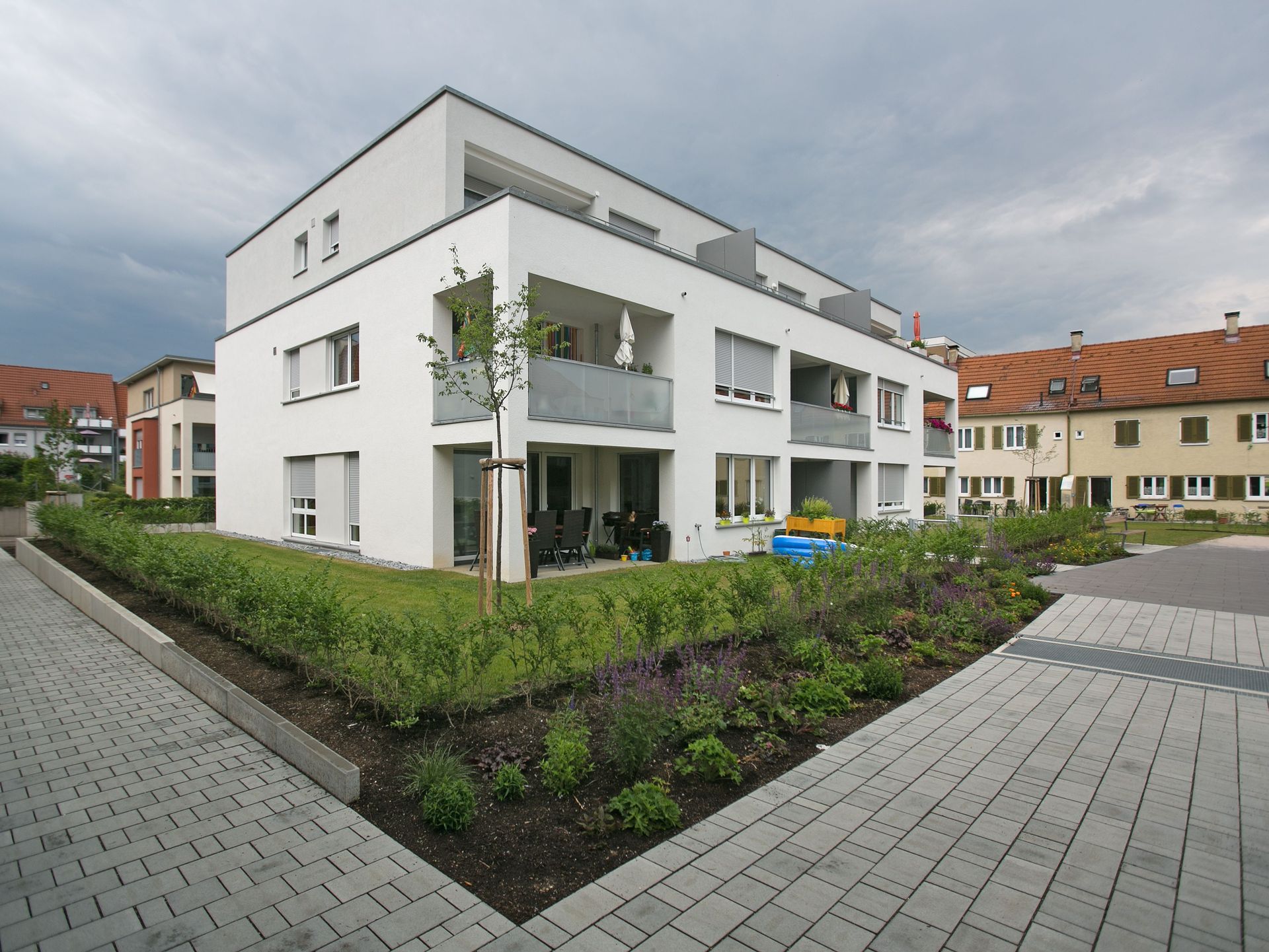 Das Bild zeigt ein modernes Mehrfamilienhaus mit weißer Fassade. Vor dem Haus befindet sich ein kleiner Gartenbereich mit Rasenfläche und einem jungen Baum.