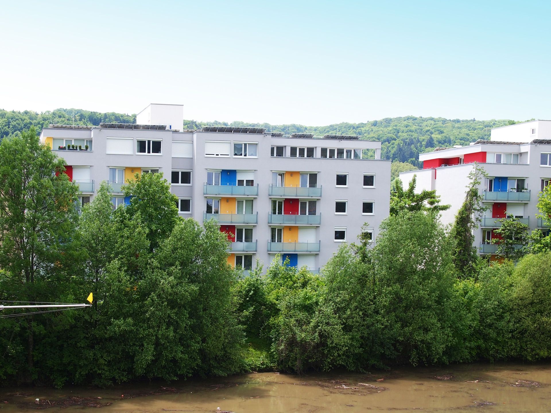 Das Bild zeigt moderne Mehrfamilienhäuser mit grauen Fassaden und Balkonen, wobei einige Türen und Fensterläden in bunten Farben wie Rot, Blau und Gelb Akzente setzen. Die Häuser stehen am Ufer eines Flusses und sind von dichtem Grün umgeben.