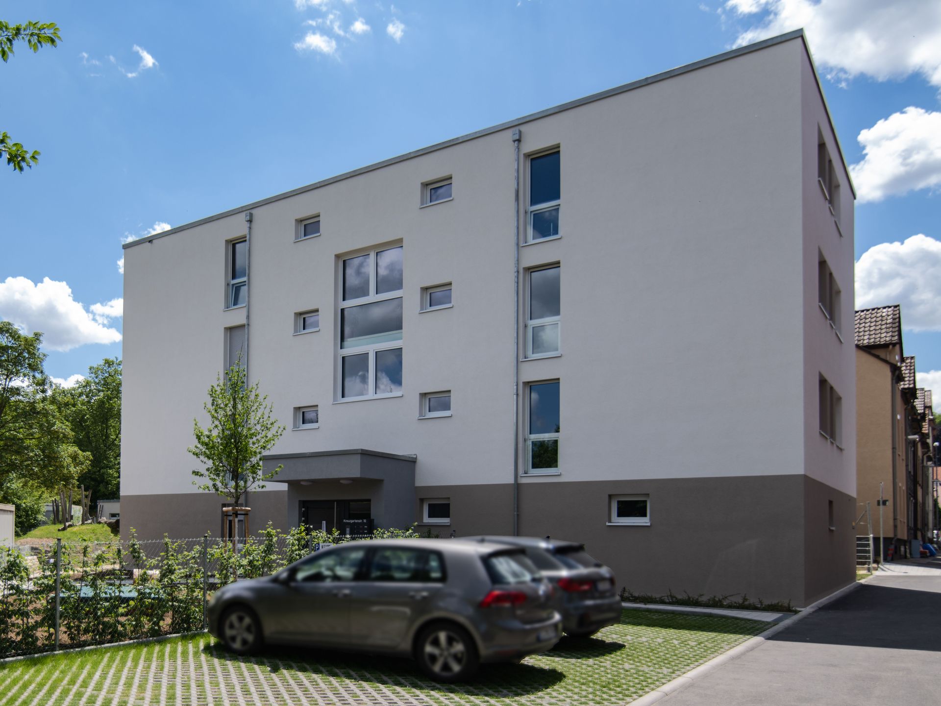 Das Bild zeigt ein modernes Mehrfamilienhaus mit weißer Fassade und grauen Akzenten. Vor dem Haus befinden sich Parkplätze und ein Zaun, dahinter ist eine Grünfläche zu erkennen.