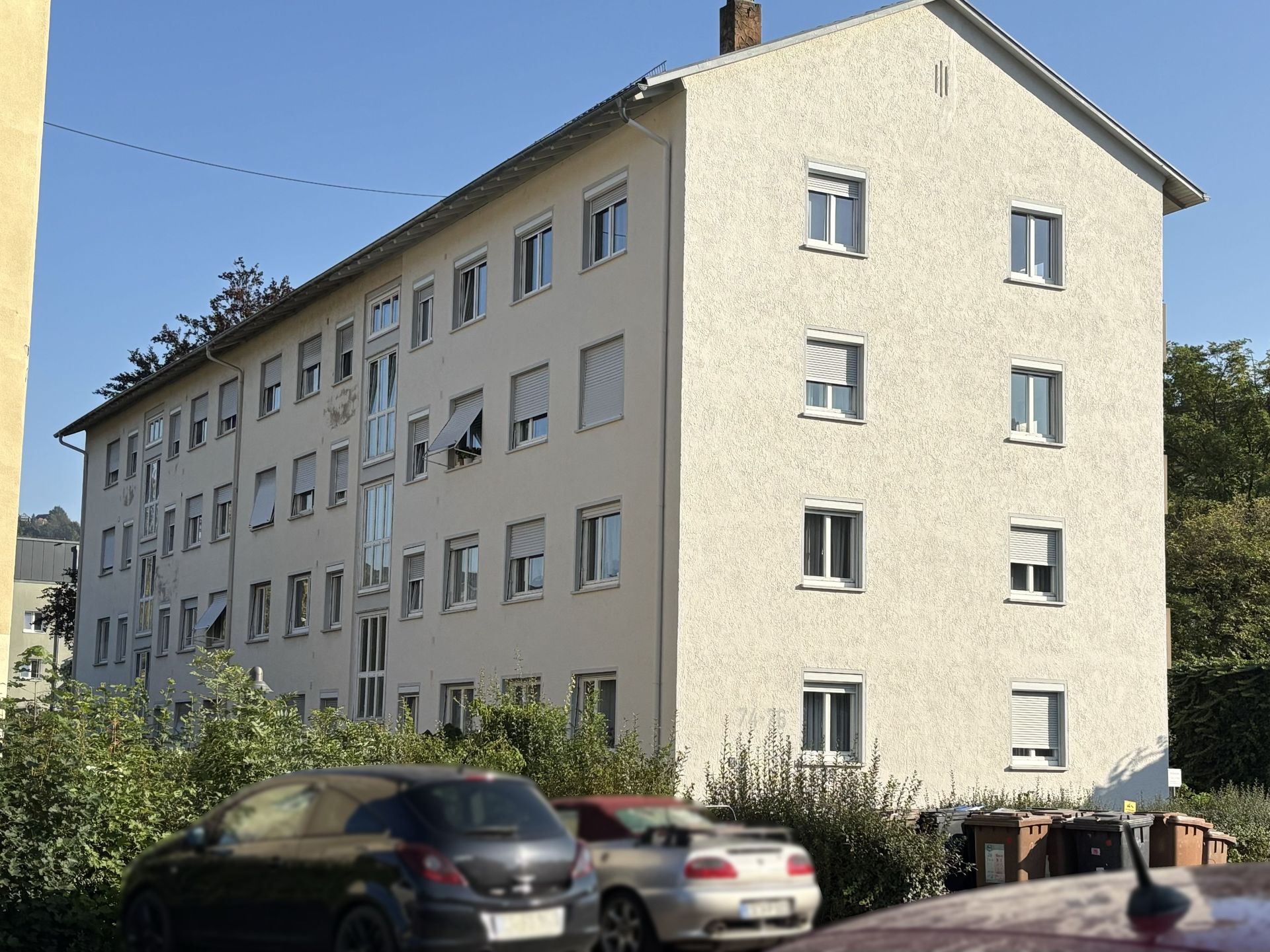 Das Bild zeigt ein Mehrfamilienhaus mit heller Fassade und vielen Fenstern. Vor dem Haus stehen mehrere Autos am Straßenrand geparkt.