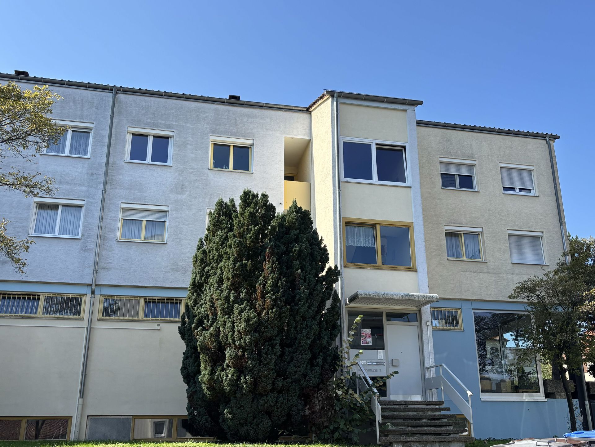 Das Bild zeigt ein Mehrfamilienhaus mit heller Fassade und mehreren Eingängen. Vor dem Haus befindet sich eine Treppe und sie führt zu einem der Eingänge.