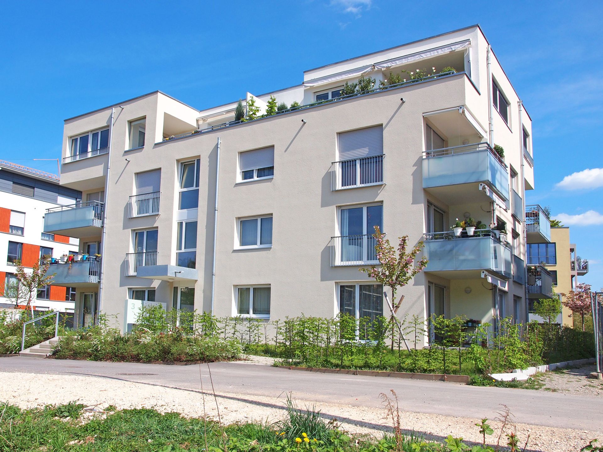 Das Bild zeigt ein Mehrfamilienhaus mit heller Fassade und Balkonen. Vor dem Haus befindet sich eine kleine Hecke.