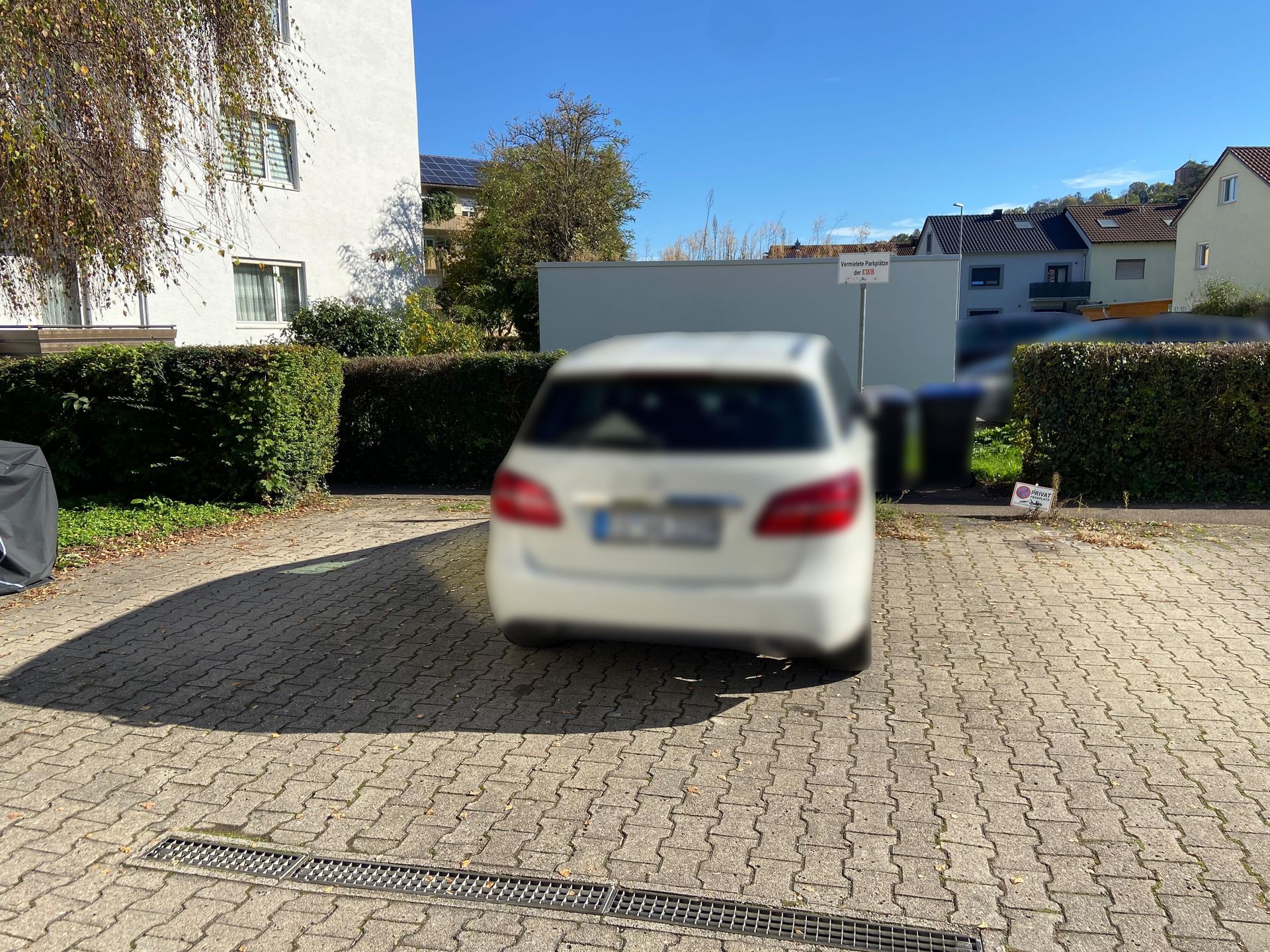 Das Bild zeigt einen Parkplatz mit einem geparkten Auto. Der Parkplatz ist von einer Hecke umgeben und im Hintergrund sind weitere Gebäude zu sehen.