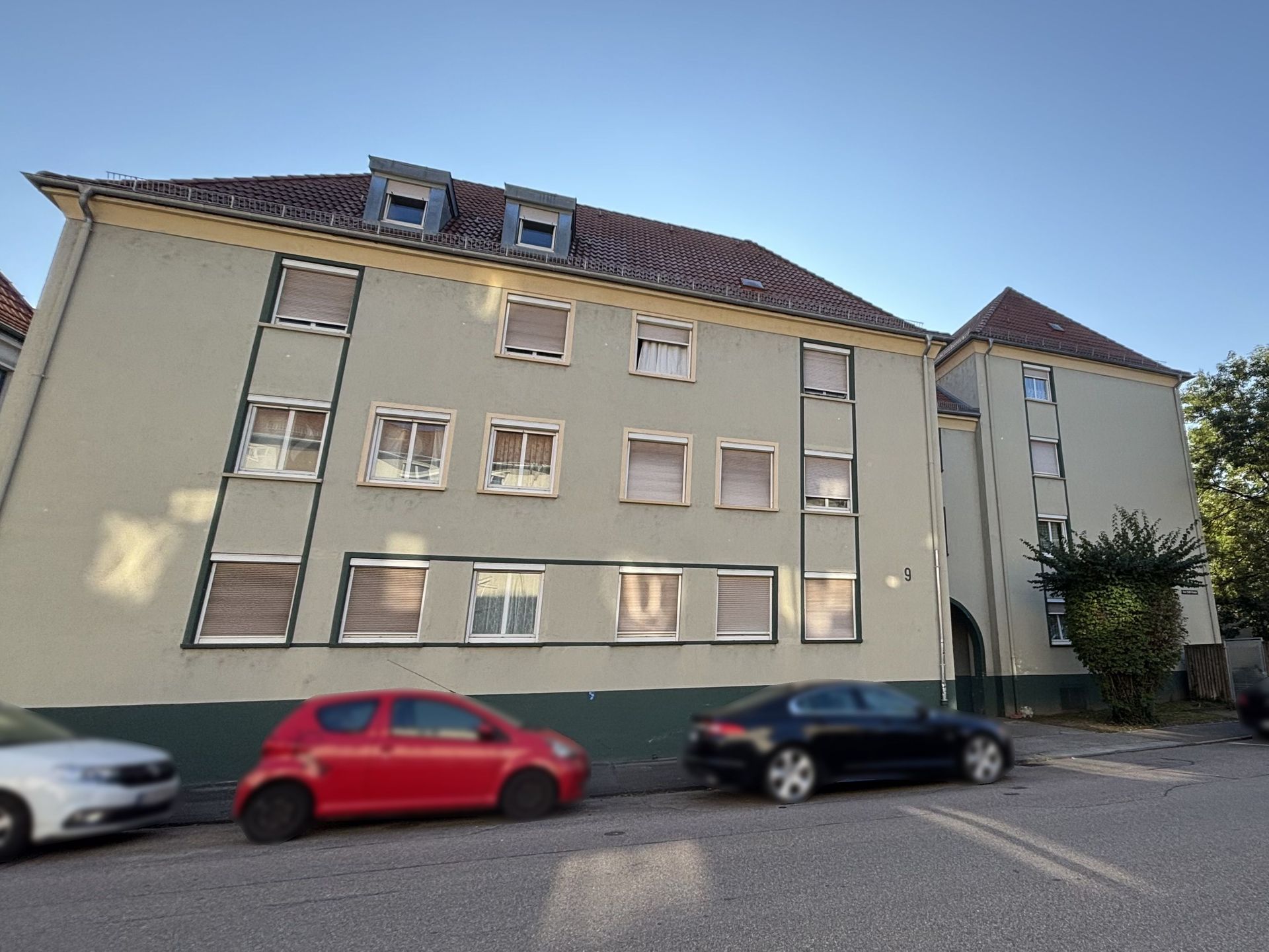 Das Bild zeigt ein Mehrfamilienhaus mit hellgrüner Fassade und dunklen Fensterrahmen. Vor dem Haus parken zwei Autos am Straßenrand.