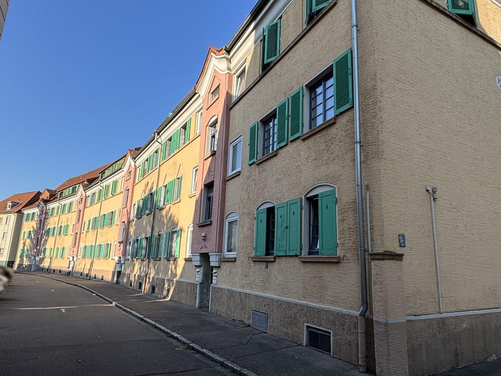 Das Bild zeigt eine Reihe von aneinandergereihten Mehrfamilienhäusern mit hellgelber Fassade und grünen Fensterläden. Die Häuser stehen an einer leicht geschwungenen Straße.
