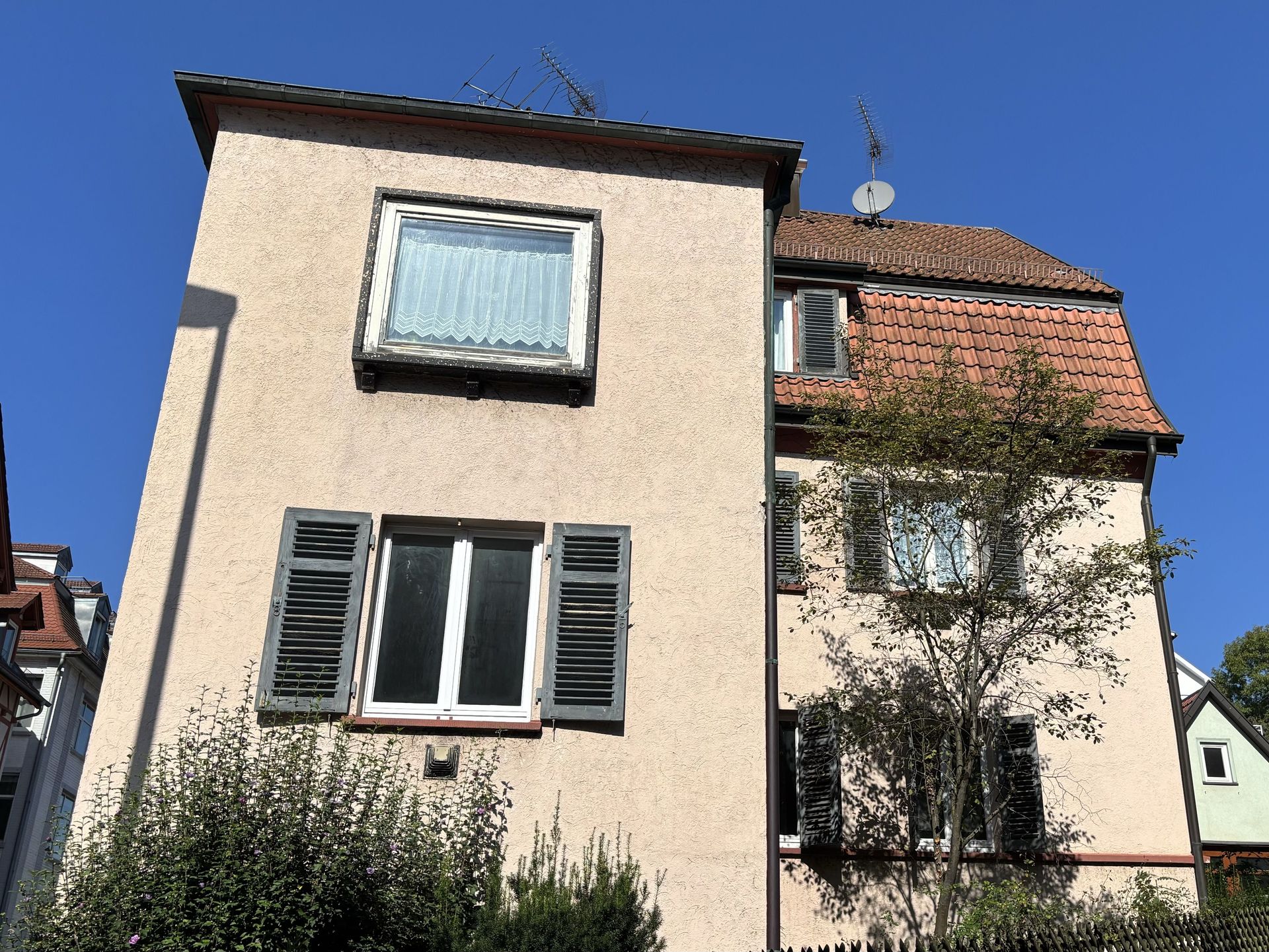 Das Bild zeigt ein helles Mehrfamilienhaus mit Satellitenschüssel und Fensterläden. Das Haus hat ein teilweise rotes Ziegeldach und ist von Bäumen und Sträuchern umgeben.