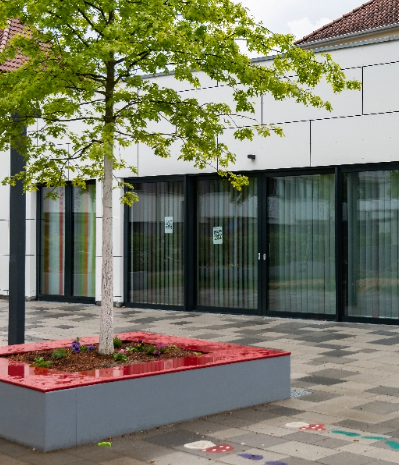 Das Bild zeigt eine Hausfassade mit großen Fensterelementen. Vorne im Bild sieht man einen Baum der von einer Sitzfläche umgeben ist. Link zum Bereich Gemeinwesenarbeit Brühl.