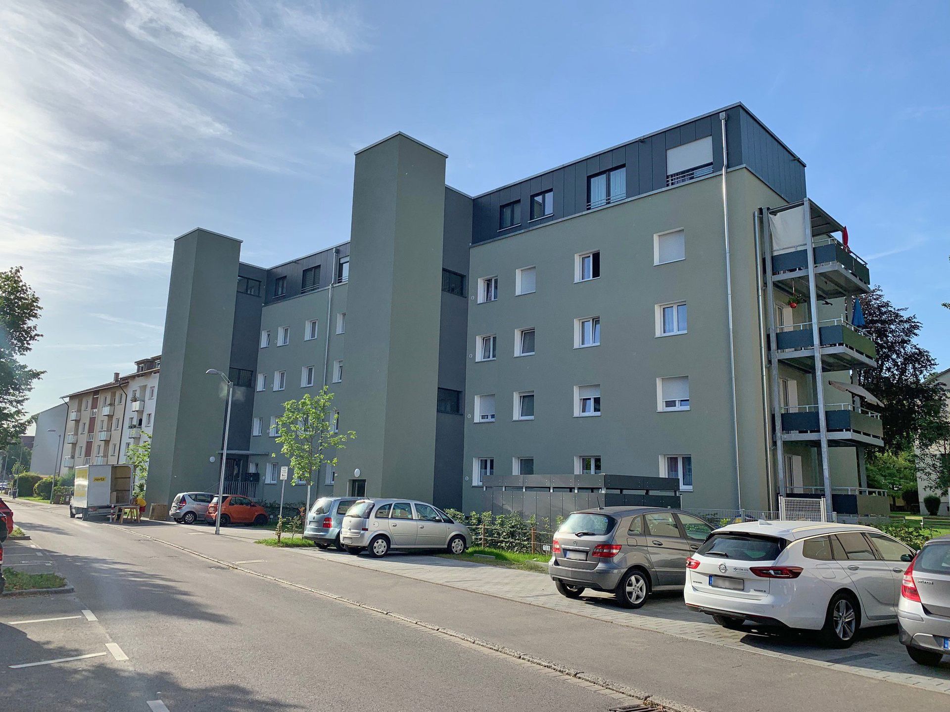 Das Bild zeigt ein Mehrfamilienhaus. Vor dem Haus sind Parkplätze auf denen Autos parken.
