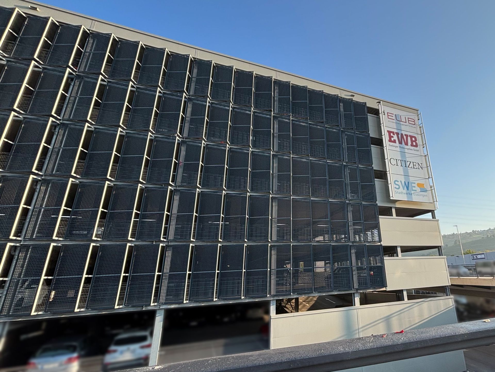 Das Bild zeigt ein Parkhaus mit einer Fassade aus hellem Beton und einer auffälligen, gitterartigen Verkleidung aus dunklem Metall. An der Seite des Parkhauses ist ein großes Banner mit dem Logo der EWB angebracht.