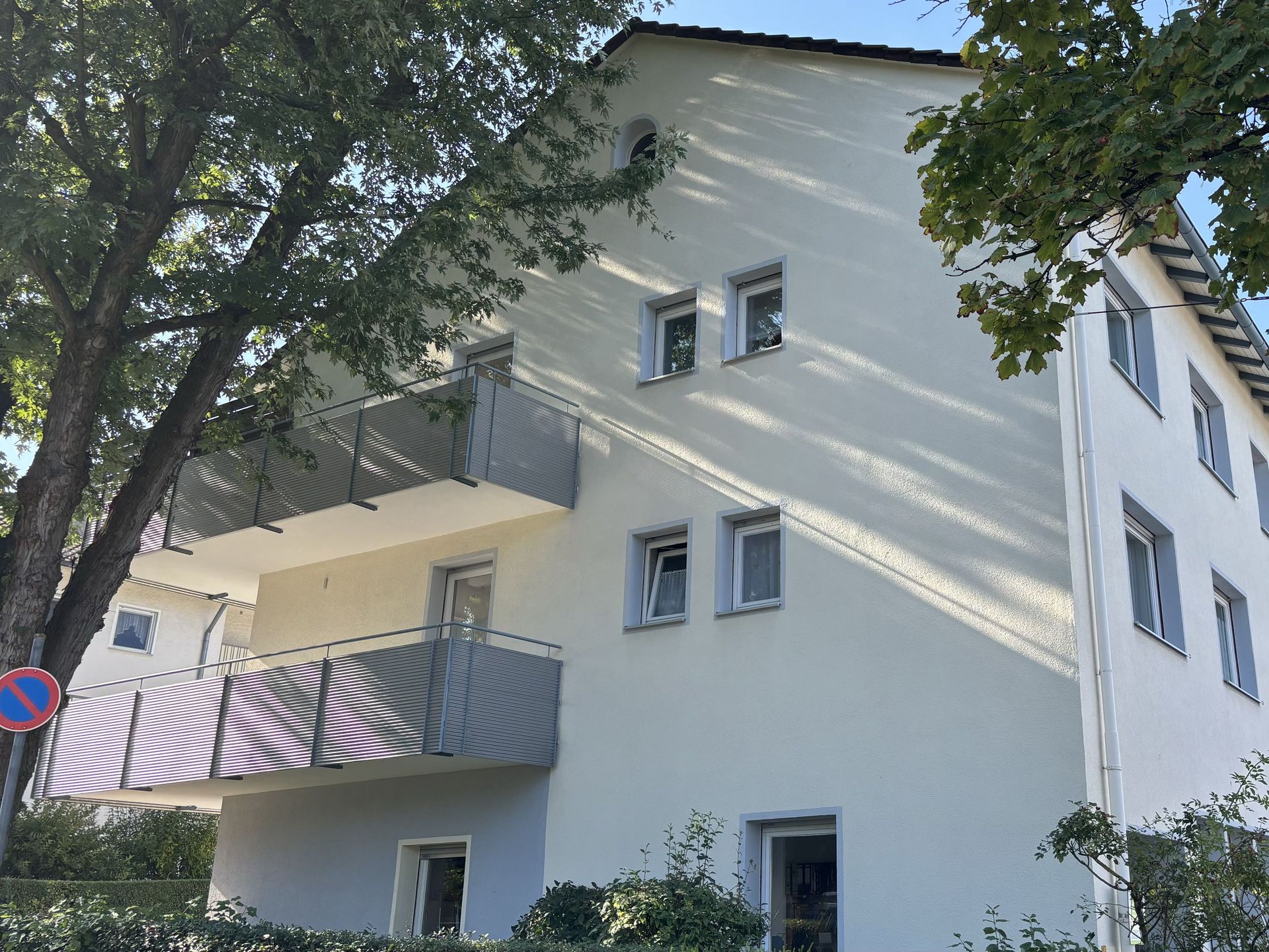 Das Bild zeigt ein Mehrfamilienhaus mit heller Fassade und Balkonen. Das Haus steht an einer Straße und ist teilweise von Bäumen verdeckt.