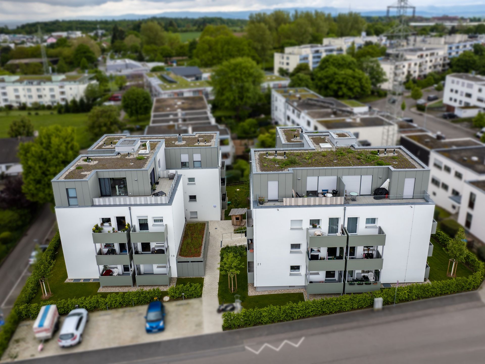 Das Bild zeigt eine moderne Wohnanlage mit begrünten Dächern und weißen Fassaden, die von viel Grün umgeben ist. Die Anlage besteht aus mehreren Gebäuden mit Balkonen und Terrassen, zwischen denen sich gepflegte Wege und kleine Gärten befinden.