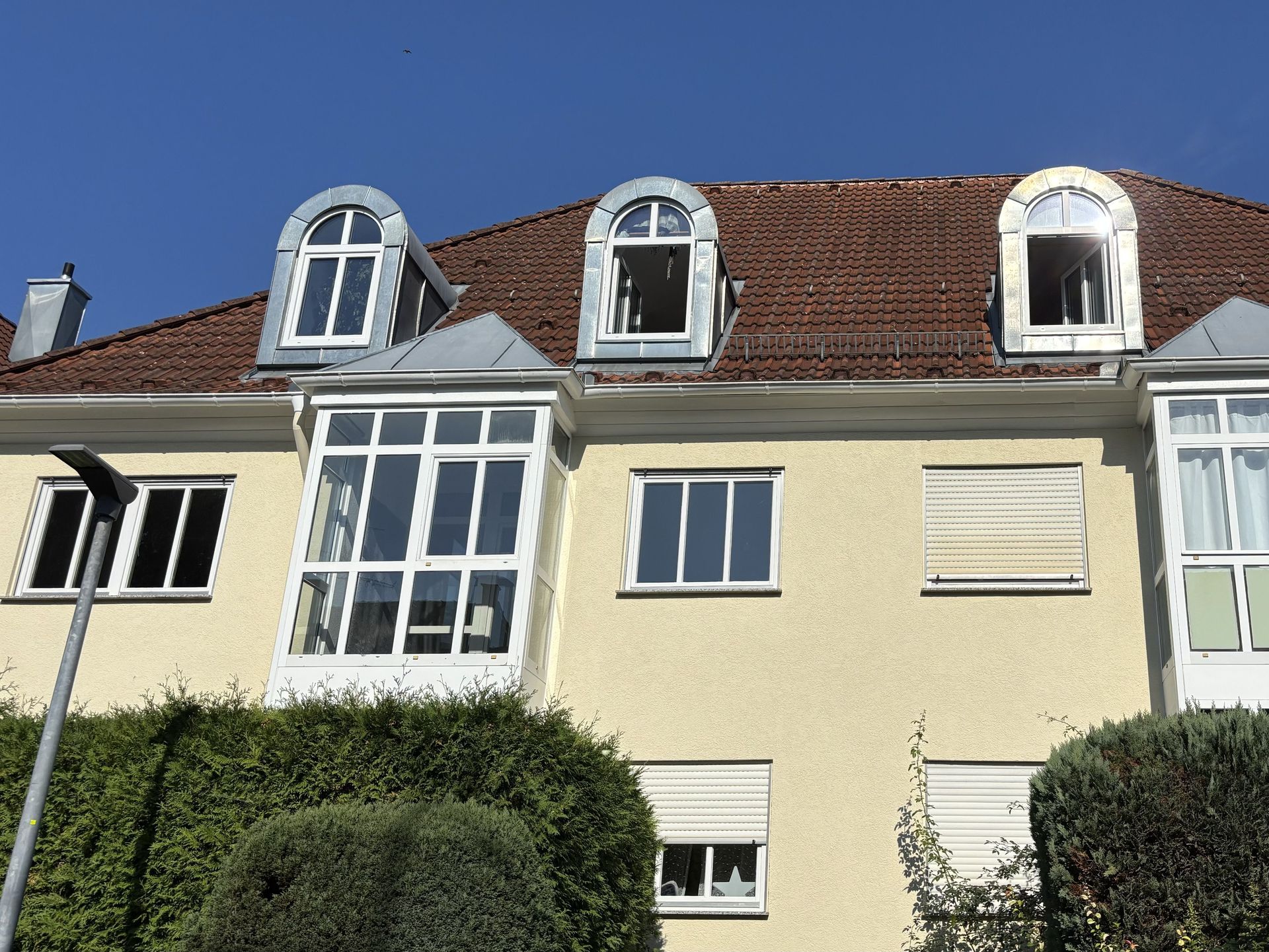Das Bild zeigt ein Mehrfamilienhaus mit hellgelber Fassade und einem roten Ziegeldach mit auffälligen, halbrunden Gauben.  Vor dem Haus befindet sich eine grüne Hecke, die die untere Etage teilweise verdeckt.
