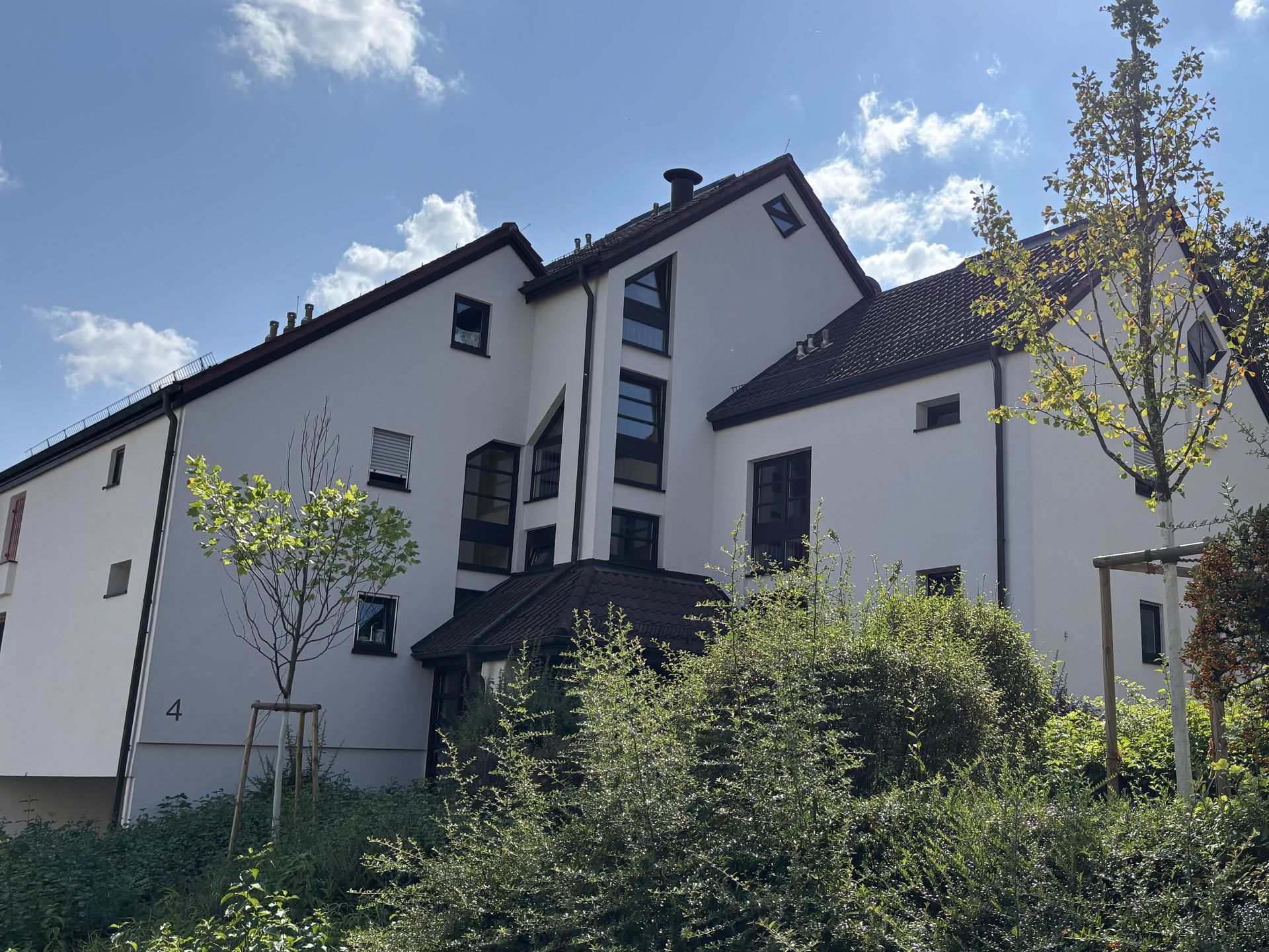 Das Bild zeigt ein modernes Mehrfamilienhaus mit weißer Fassade und dunklem Dach. Vor dem Haus befinden sich Bäume und Sträucher.