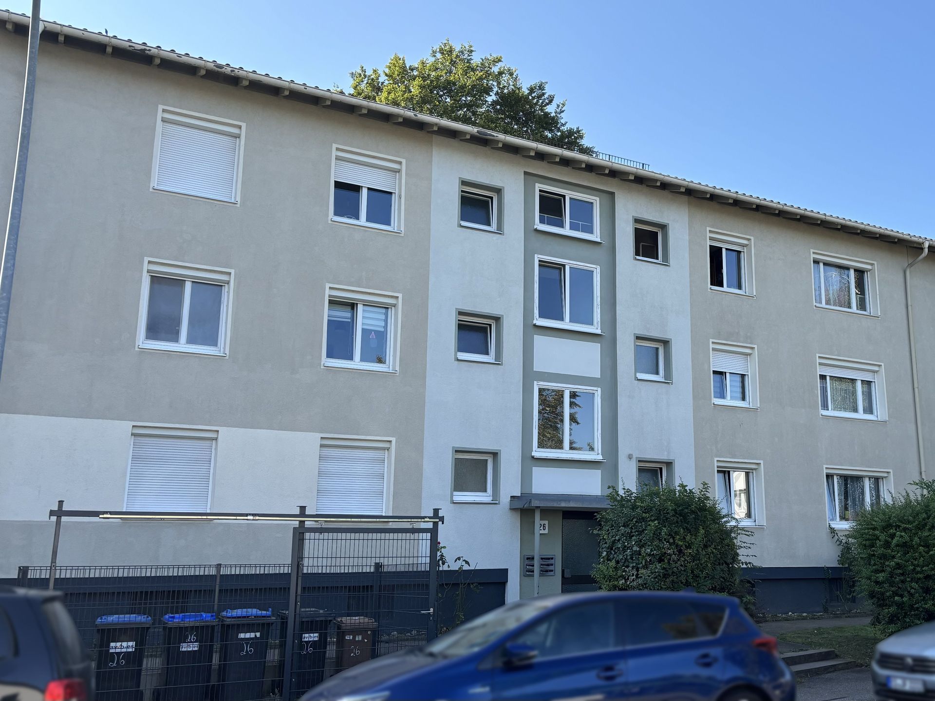 Das Bild zeigt ein Mehrfamilienhaus mit heller Fassade und weißen Fenstern. Vor dem Haus stehen Mülltonnen und ein Auto parkt am Straßenrand.