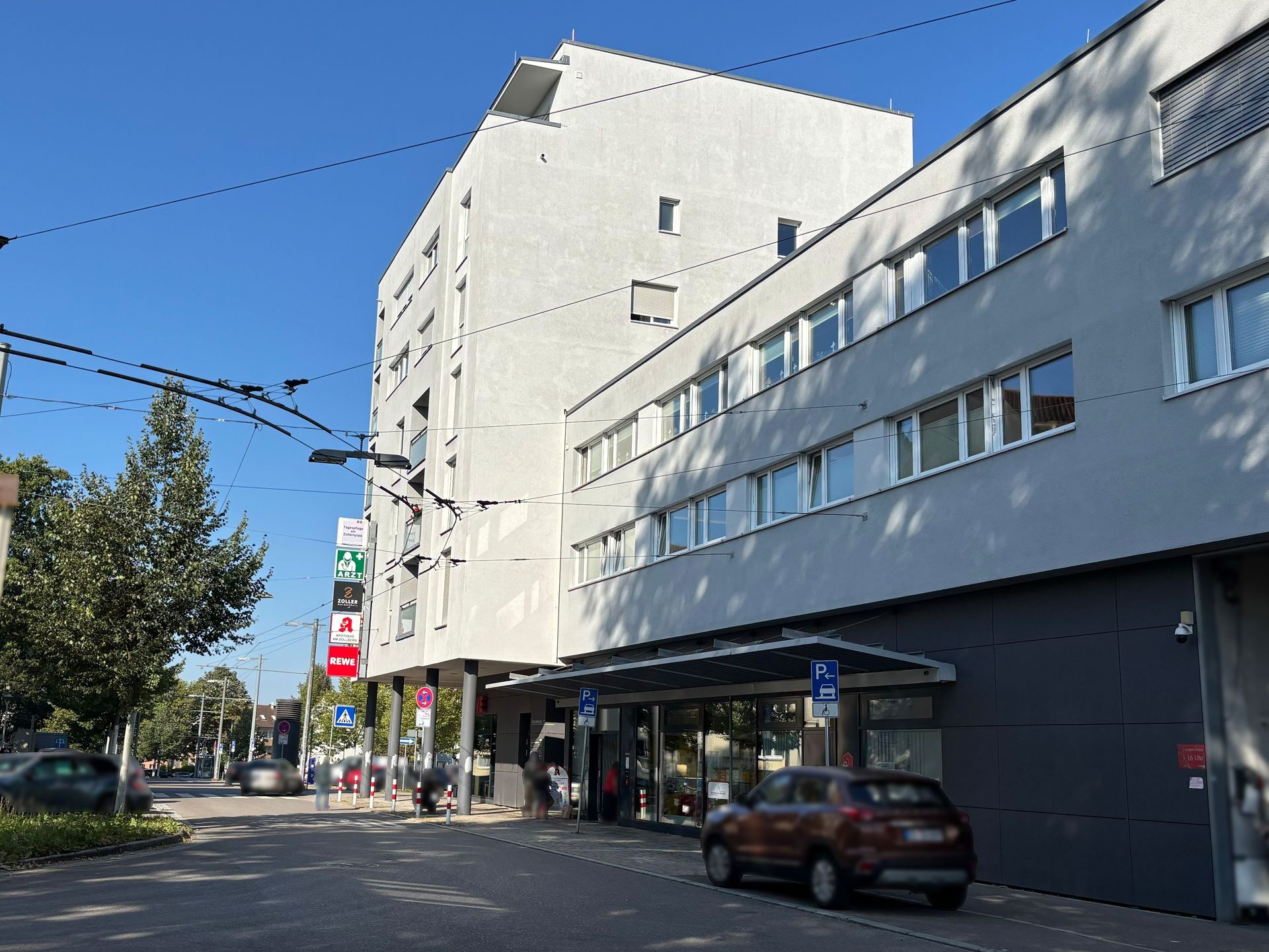 Das Bild zeigt ein modernes Gebäude an einer Straße mit Oberleitungen. Im Erdgeschoss des Gebäudes befinden sich Läden oder Geschäfte.