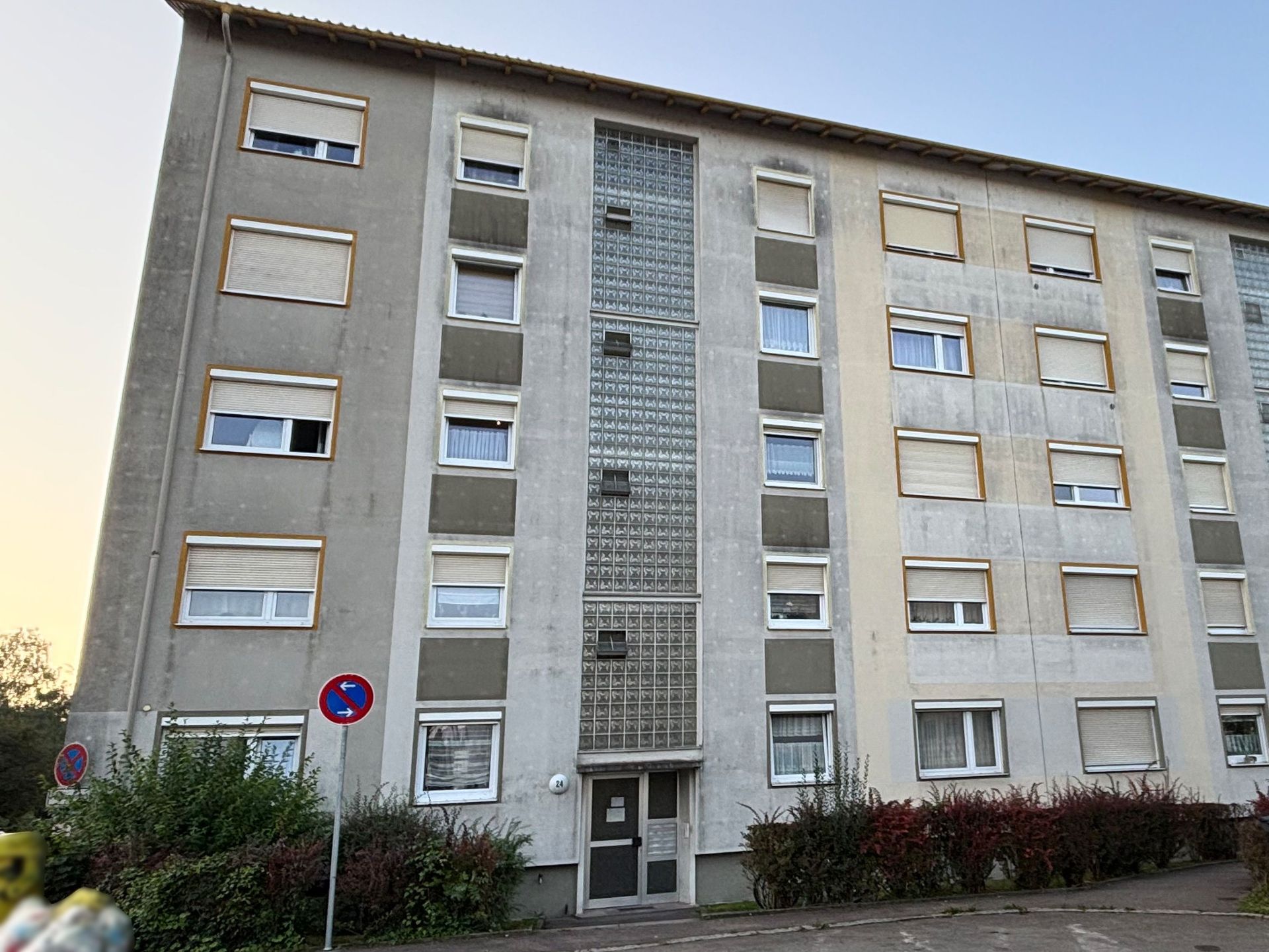 Das Bild zeigt ein Mehrfamilienhaus mit grauer Fassade und einem auffälligen vertikalen Glaseinsatz über der Eingangstür. Vor dem Haus befinden sich Sträucher.