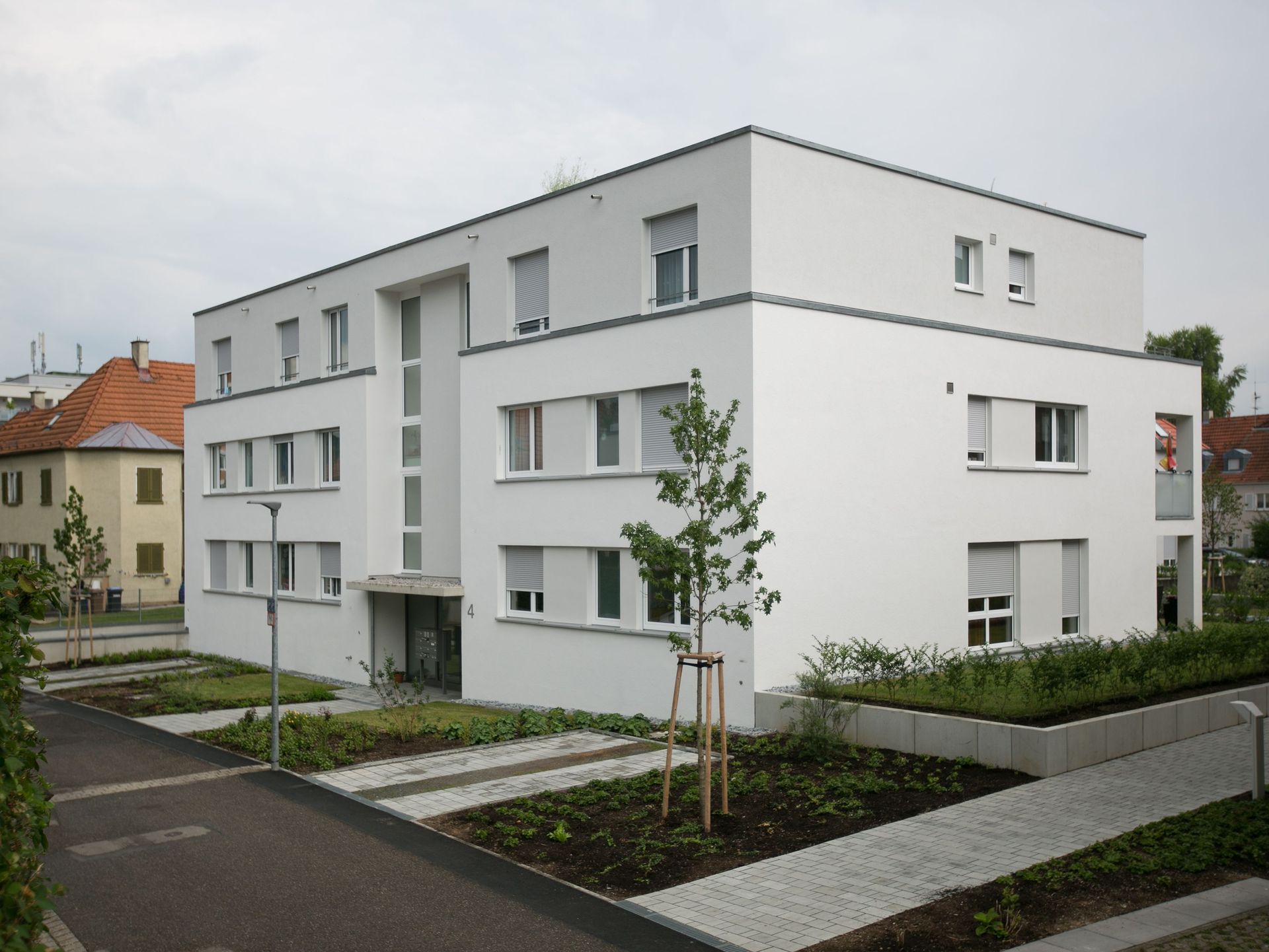 Das Bild zeigt ein modernes, kubisches Mehrfamilienhaus mit weißer Fassade.  Um das Gebäude herum sind Grünflächen, junge Bäume und gepflasterte Wege angelegt.