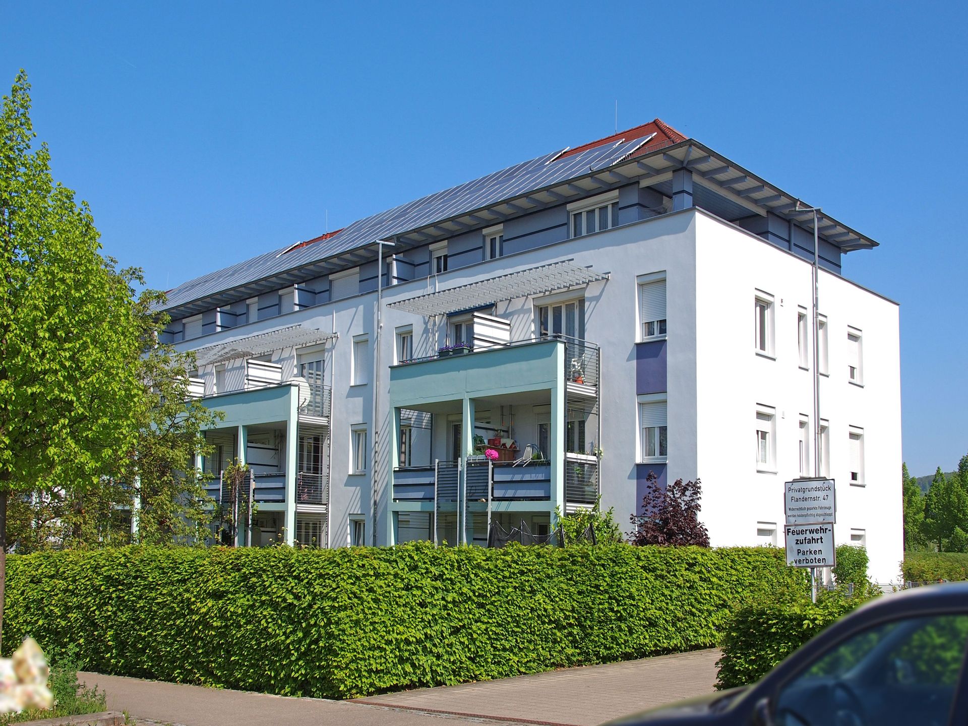 Das Bild zeigt ein modernes Mehrfamilienhaus mit weißer Fassade, das durch farbige Elemente an den Balkonen und Fensterbänken Akzente erhält. Das Haus besitzt eine Solaranlage auf em Dach und ist von einer dichten Hecke umgeben.