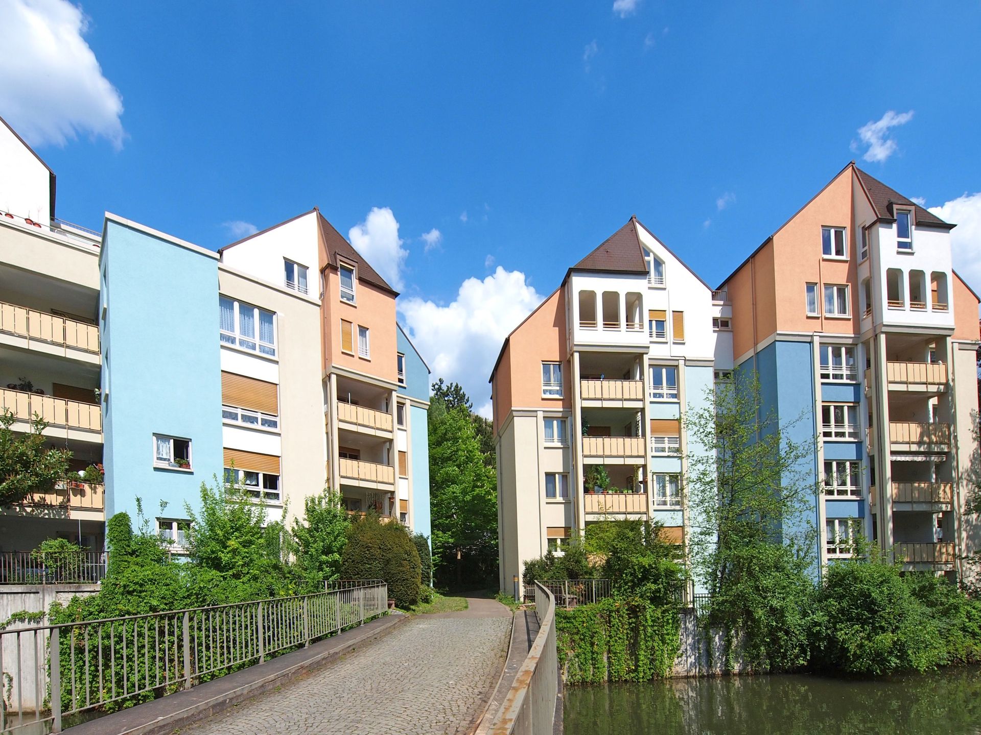 Das Bild zeigt eine Wohnanlage mit mehreren Gebäuden in verschiedenen Farben, die an einem kleinen Fluss liegen. Eine Brücke führt über das Wasser und ermöglicht den Zugang zu den Häusern, die von Bäumen und Sträuchern umgeben sind.