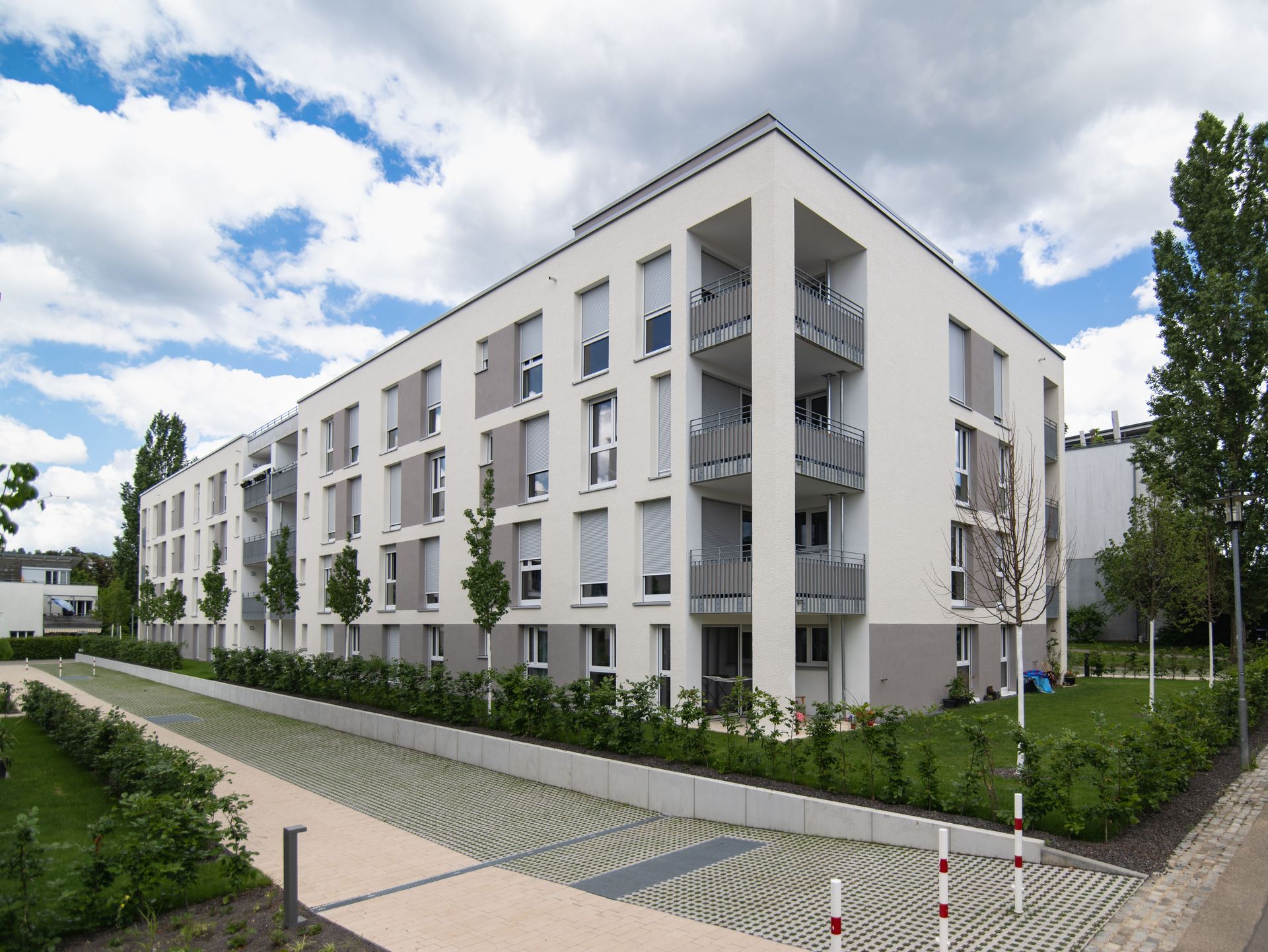 Das Bild zeigt ein Mehrfamilienhaus mit weißer Fassade. Vor dem Haus ist eine Grünfläche, die von einer kleinen Hecke umgeben ist. Davor befindet sich ein gepflasteter Weg.