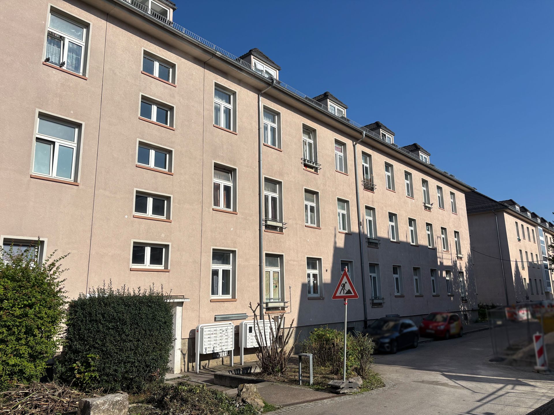 Das Bild zeigt ein helles Mehrfamilienhaus mit vielen gleichförmigen Fenstern. Vor dem Haus befindet sich ein gepflasterter Bereich mit Briefkästen, Sträuchern.