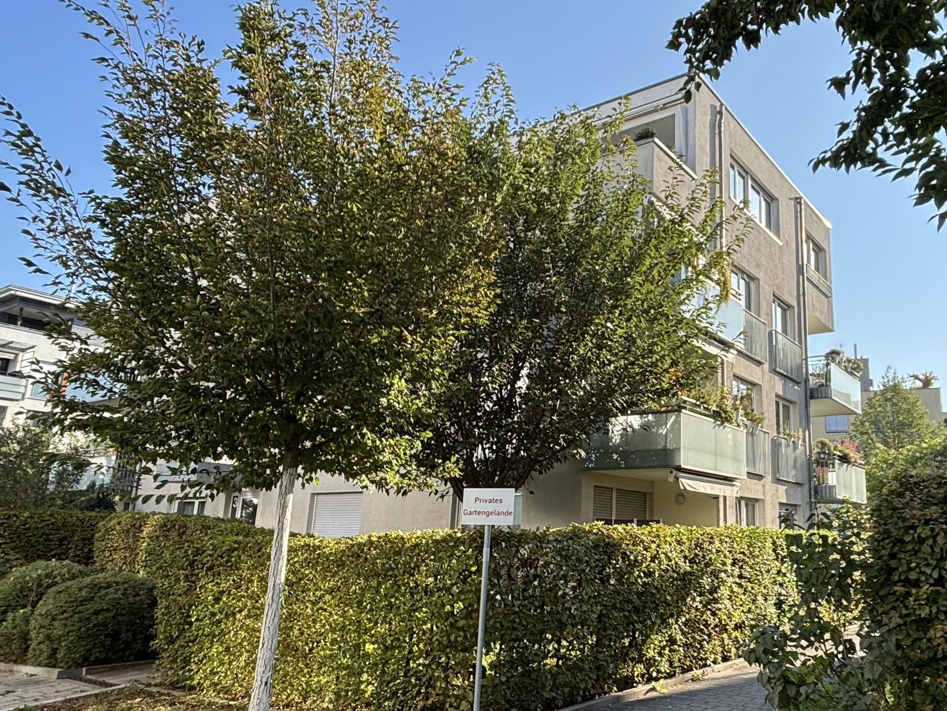 Das Bild zeigt ein modernes Mehrfamilienhaus mit heller Fassade und Balkonen. Vor dem Haus befinden sich eine dichte Hecke und zwei Bäume.
