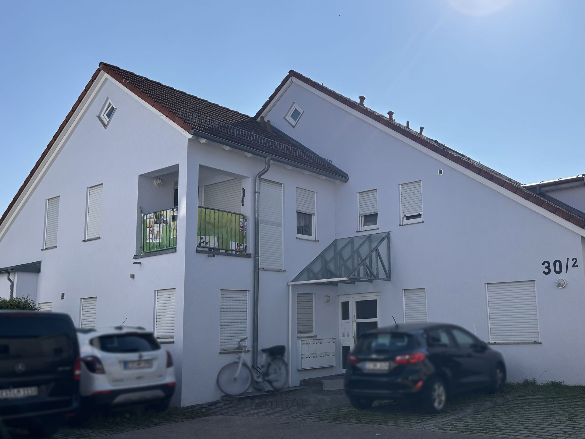 Das Bild zeigt ein Mehrfamilienhaus mit heller Fassade und einem kleinen Balkon. Vor dem Haus befinden sich Parkplätze, auf denen Autos abgestellt sind.