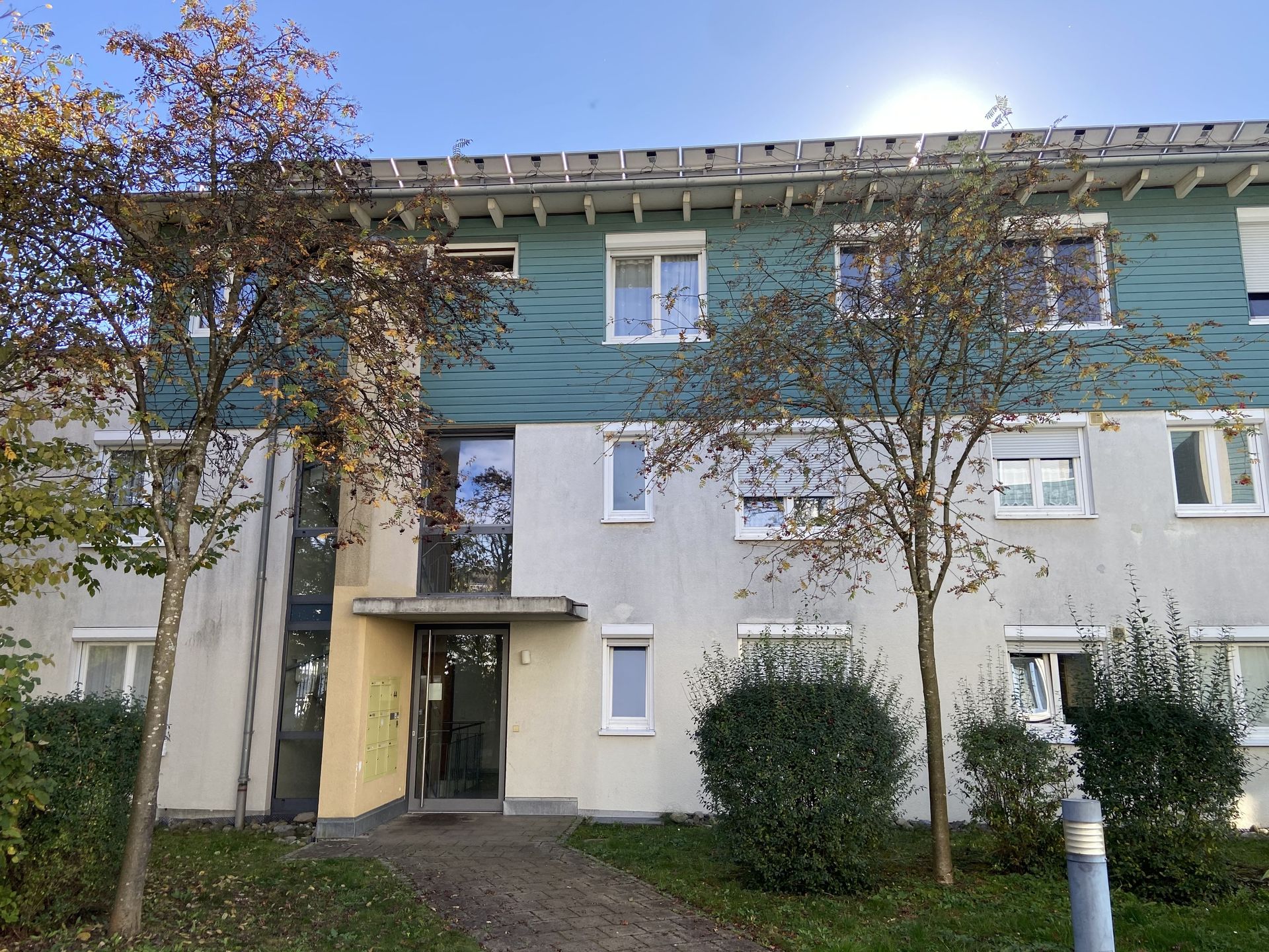 Das Bild zeigt ein Mehrfamilienhaus mit grauer Fassade und einem auffälligen, türkisfarbenen Holzelement im oberen Bereich. Vor dem Haus befinden sich Bäume, Sträucher und ein gepflasterter Weg, der zum Eingang führt.
