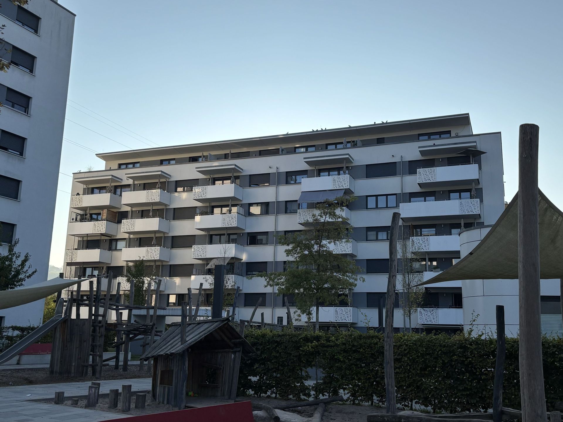 Das Bild zeigt ein modernes Mehrfamilienhaus mit weißer Fassade, Balkonen und großen Fenstern. Im Vordergrund befindet sich ein Spielplatz mit Klettergerüst und Rutsche, der von einer Hecke umgeben ist.