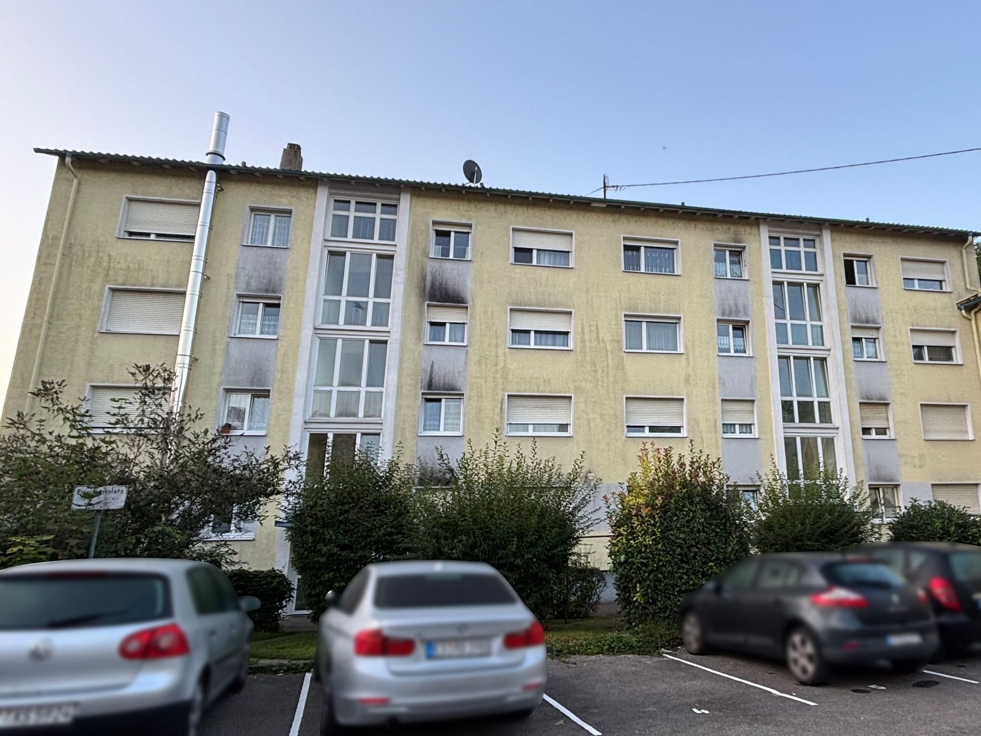 Das Bild zeigt ein Mehrfamilienhaus mit hellgelber Fassade. Vor dem Haus befinden sich Parkplätze, auf denen mehrere Autos abgestellt sind.