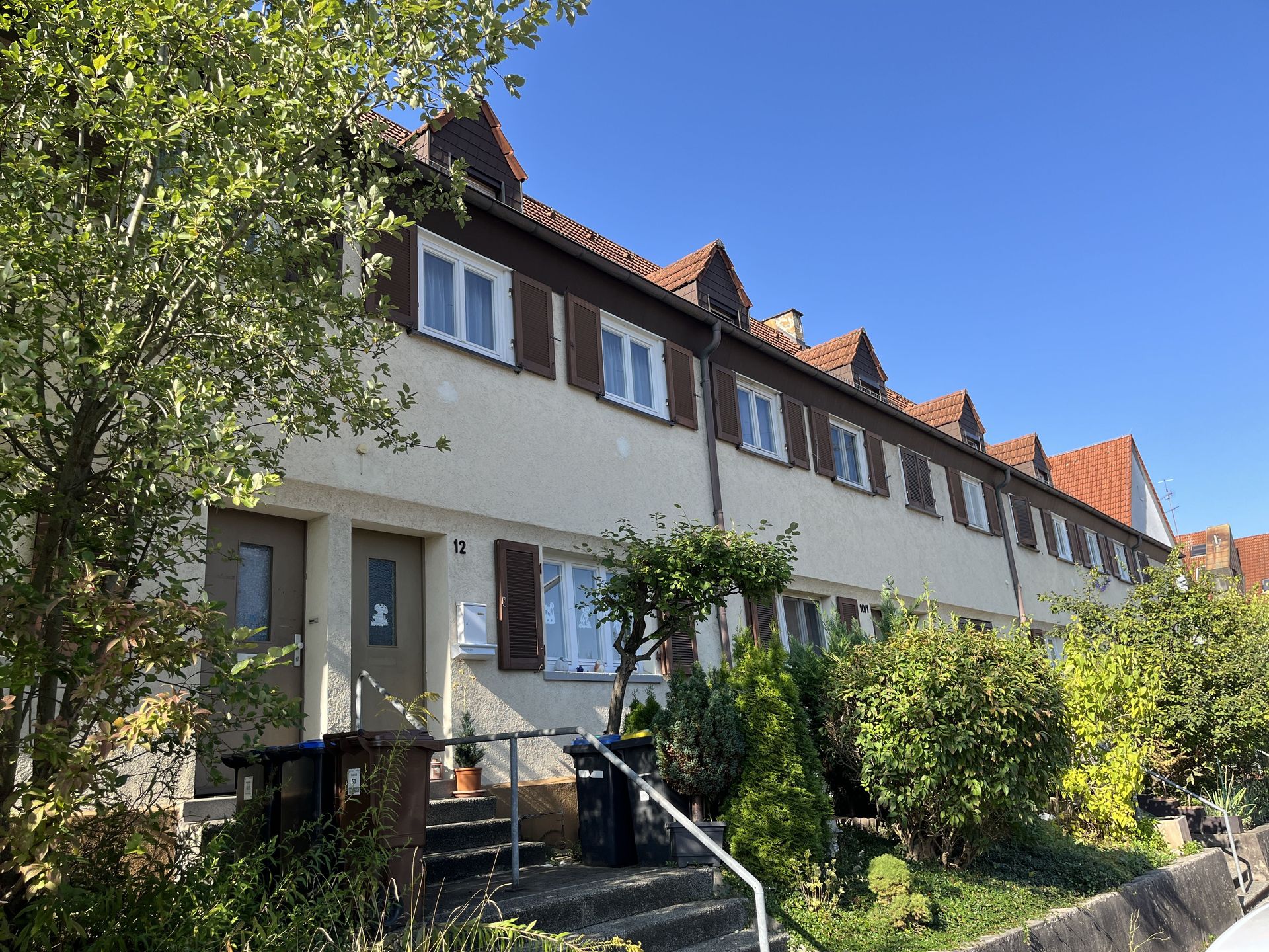 Das Bild zeigt eine Reihe von Häusern mit heller Fassade, braunen Fensterläden und roten Ziegeldächern. Vor den Häusern befinden sich kleine Vorgärten mit Sträuchern und Bäumen.