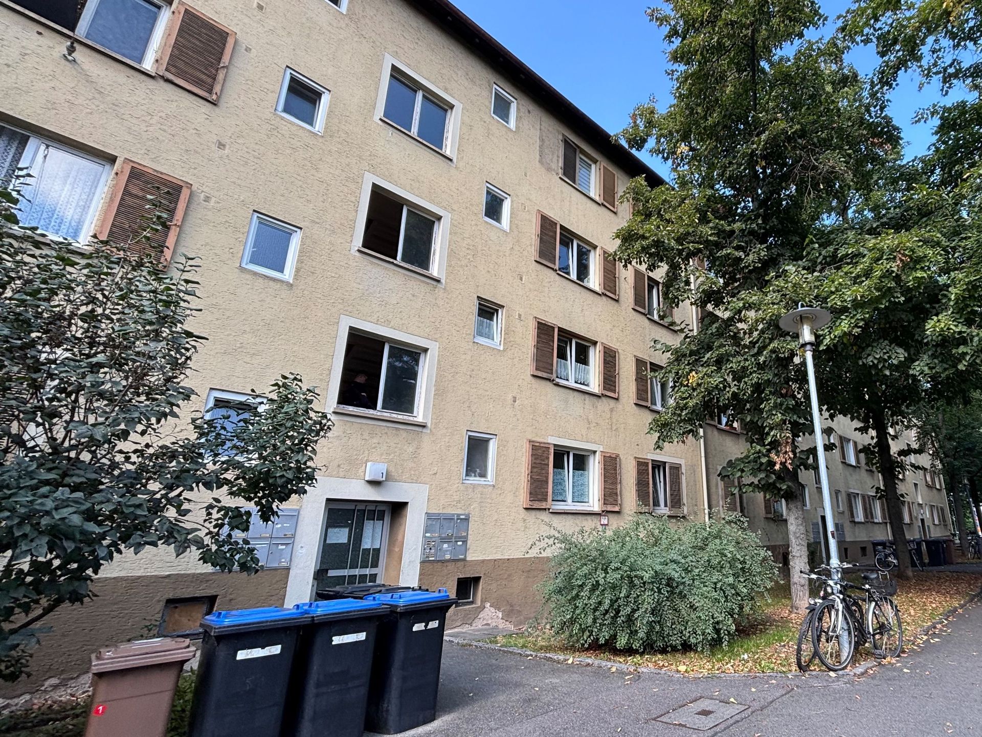 Das Bild zeigt ein Mehrfamilienhaus mit heller Fassade und braunen Fensterläden. Neben dem Haus führt ein Weg entlang, an dem Fahrräder abgestellt sind und Bäume Schatten spenden.