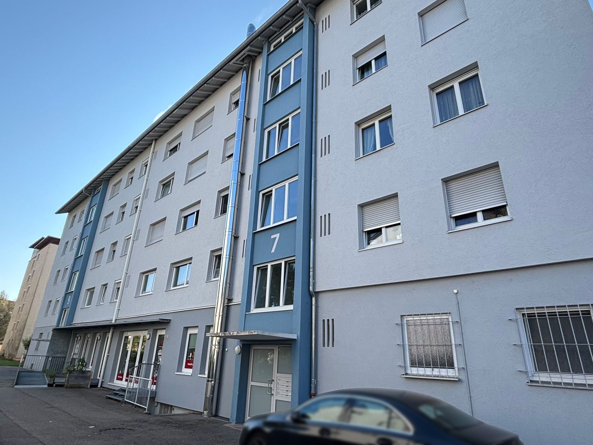 Das Bild zeigt ein Mehrfamilienhaus mit grauer Fassade und blauen Akzenten. Im Erdgeschoss befinden sich Gewerberäume, und davor parkt ein schwarzes Auto.