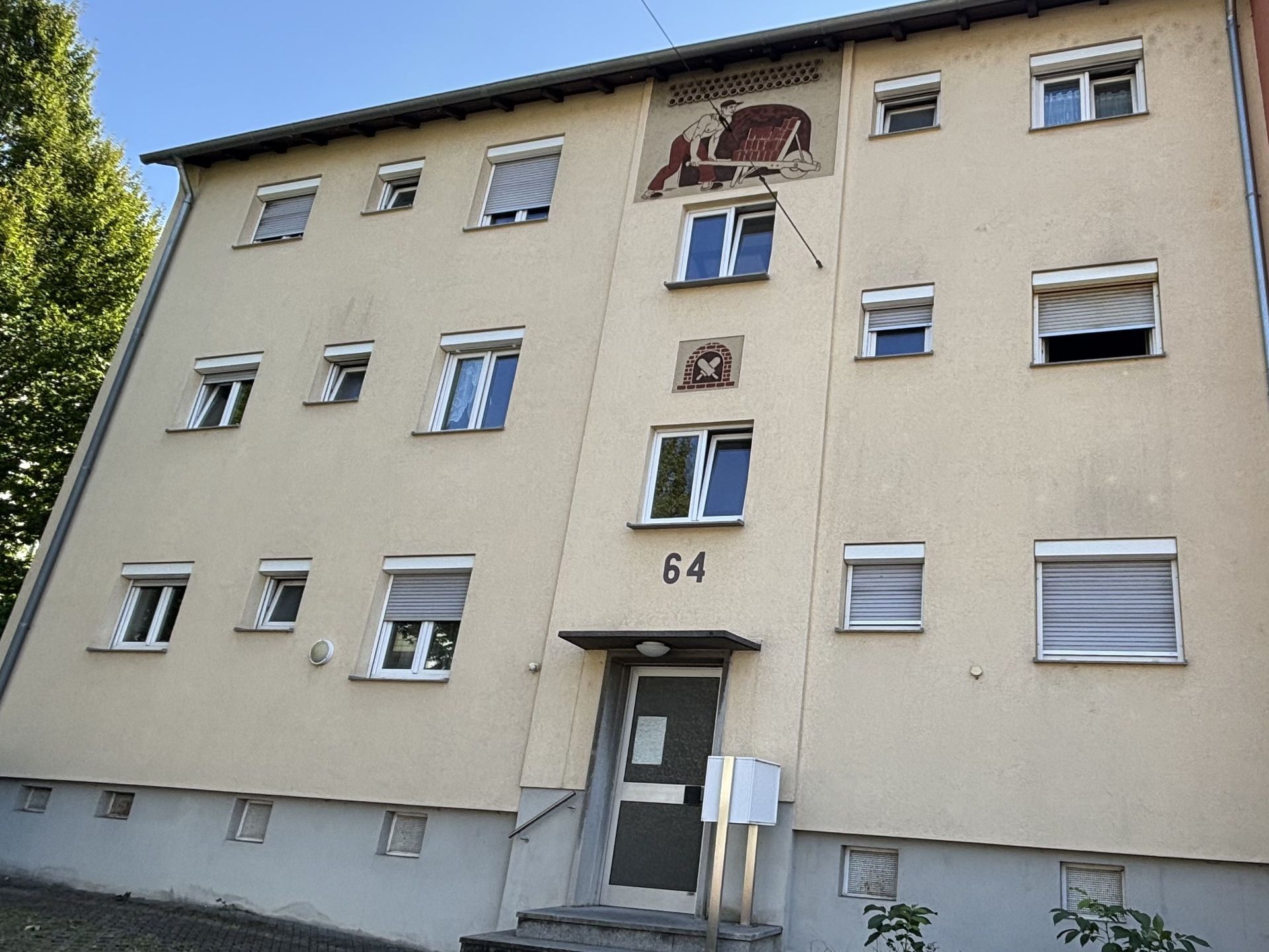 Das Bild zeigt ein Mehrfamilienhaus mit hellgelber Fassade und unterschiedlich angeordneten Fenstern. Zwischen den Fenstern über der Eingangstür schmücken Abbildungen den Haus.