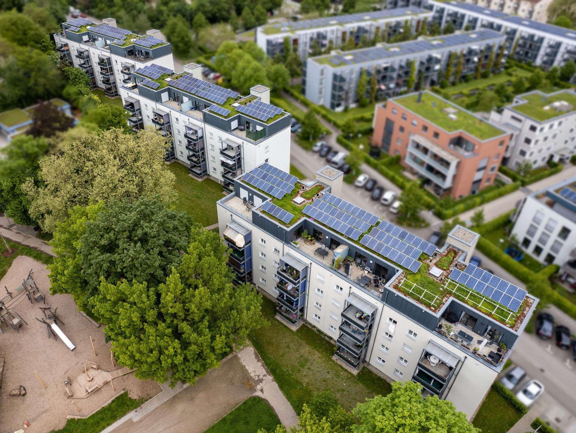 Das Bild zeigt eine Wohnanlage mit 3 Häusern. Alle Häuser haben eine begrünte Dachfläche mit Photovoltaikanlagen auf dem Dach. Die Häuser sind von Grünflächen und Bäumen umgeben.